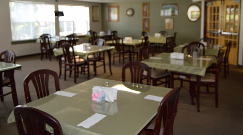 A dining room with multiple square tables covered with light green tablecloths. Each table has several dark wooden chairs around it and is set with napkins and a small centerpiece. The room has large windows letting in natural light, framed pictures on the walls, and a carpeted floor.