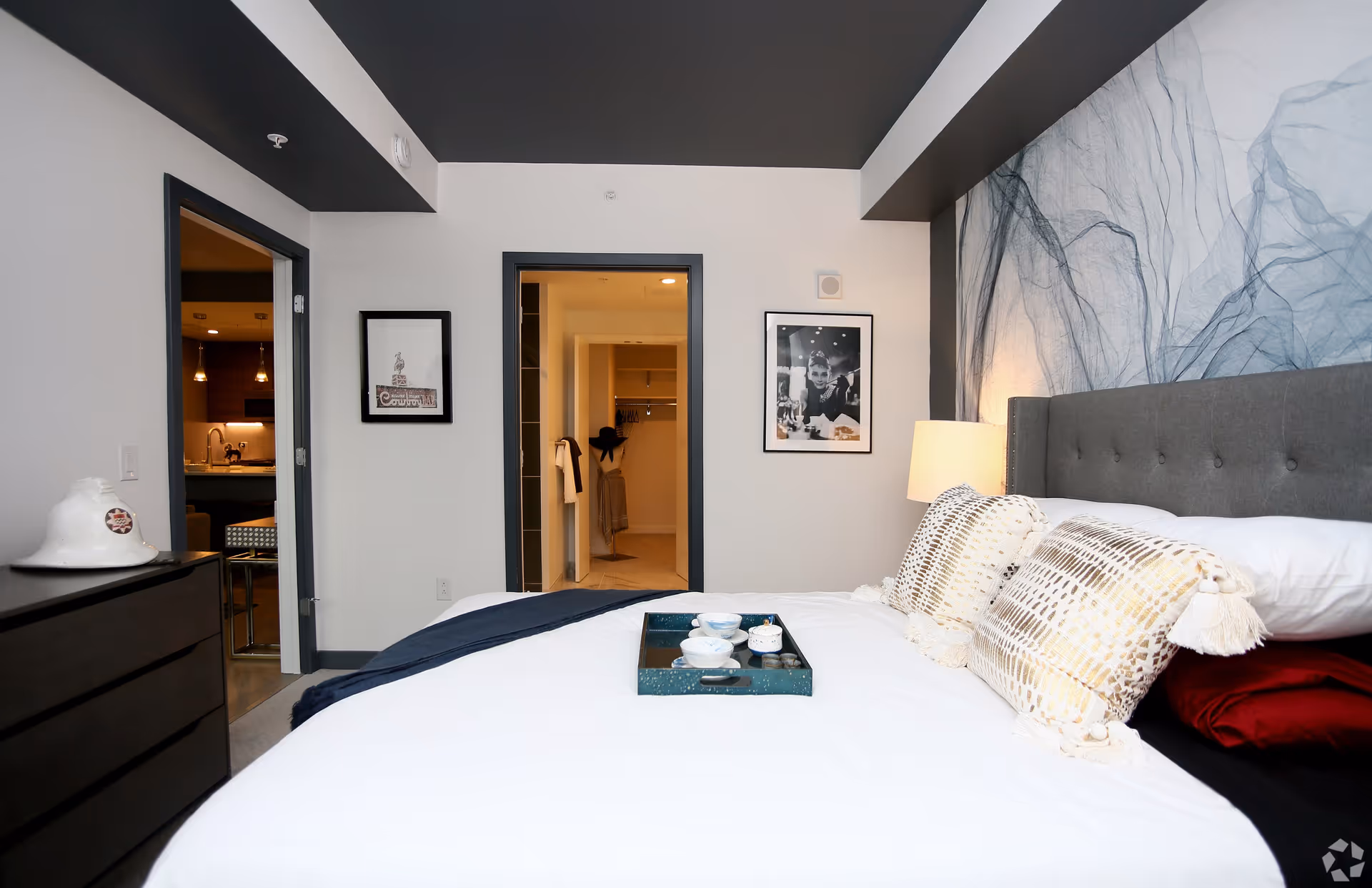 Modern bedroom with a made bed, decorative pillows and bedside lamp, and open doorways showing a bathroom and kitchen area.