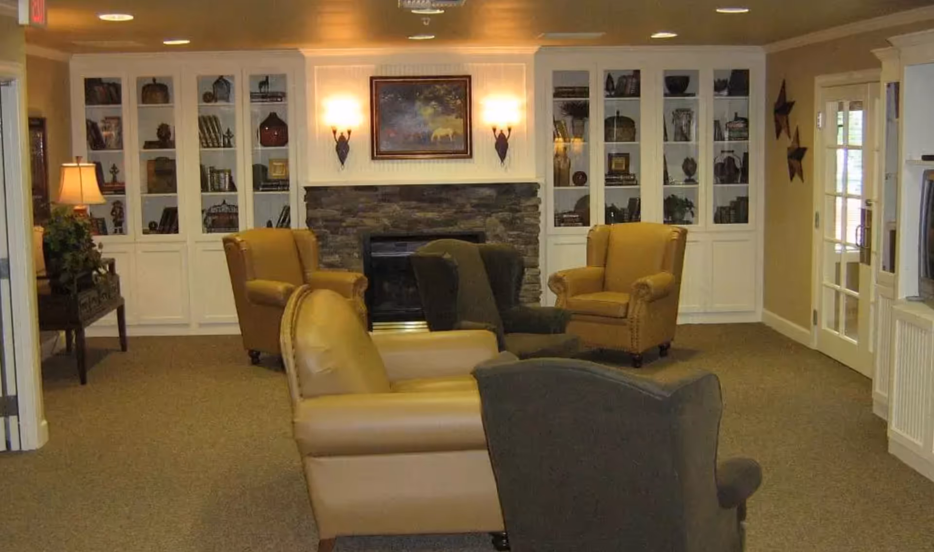 A cozy living room area with four armchairs arranged around a stone fireplace. The fireplace is flanked by white built-in cabinets filled with decorative items and books. The room is softly lit by wall sconces and a table lamp, with a framed painting above the fireplace and a glass door on the right side.