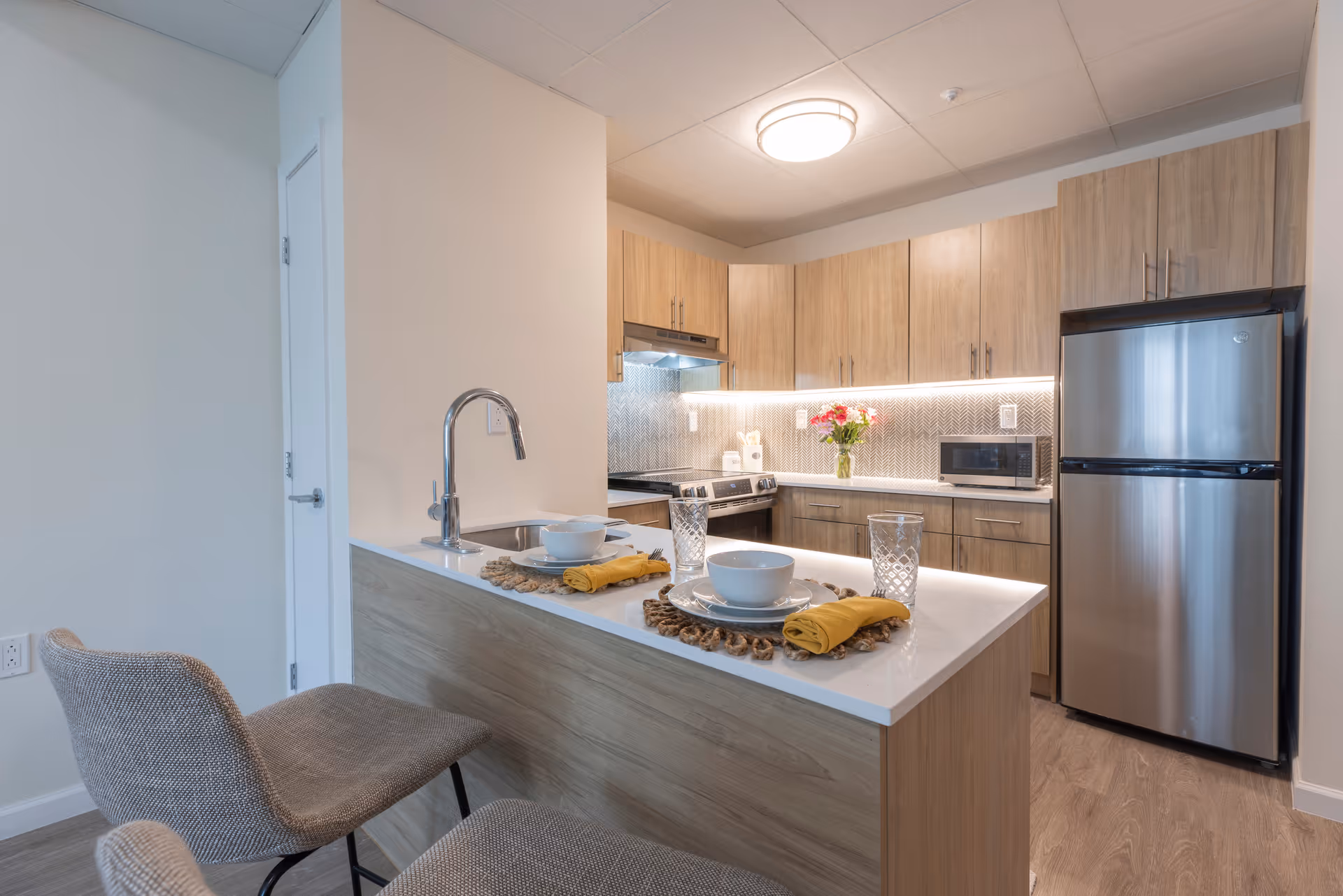 Small modern kitchen with an island and two bar stools, wood cabinets, stainless steel refrigerator, sink, stove and place settings.