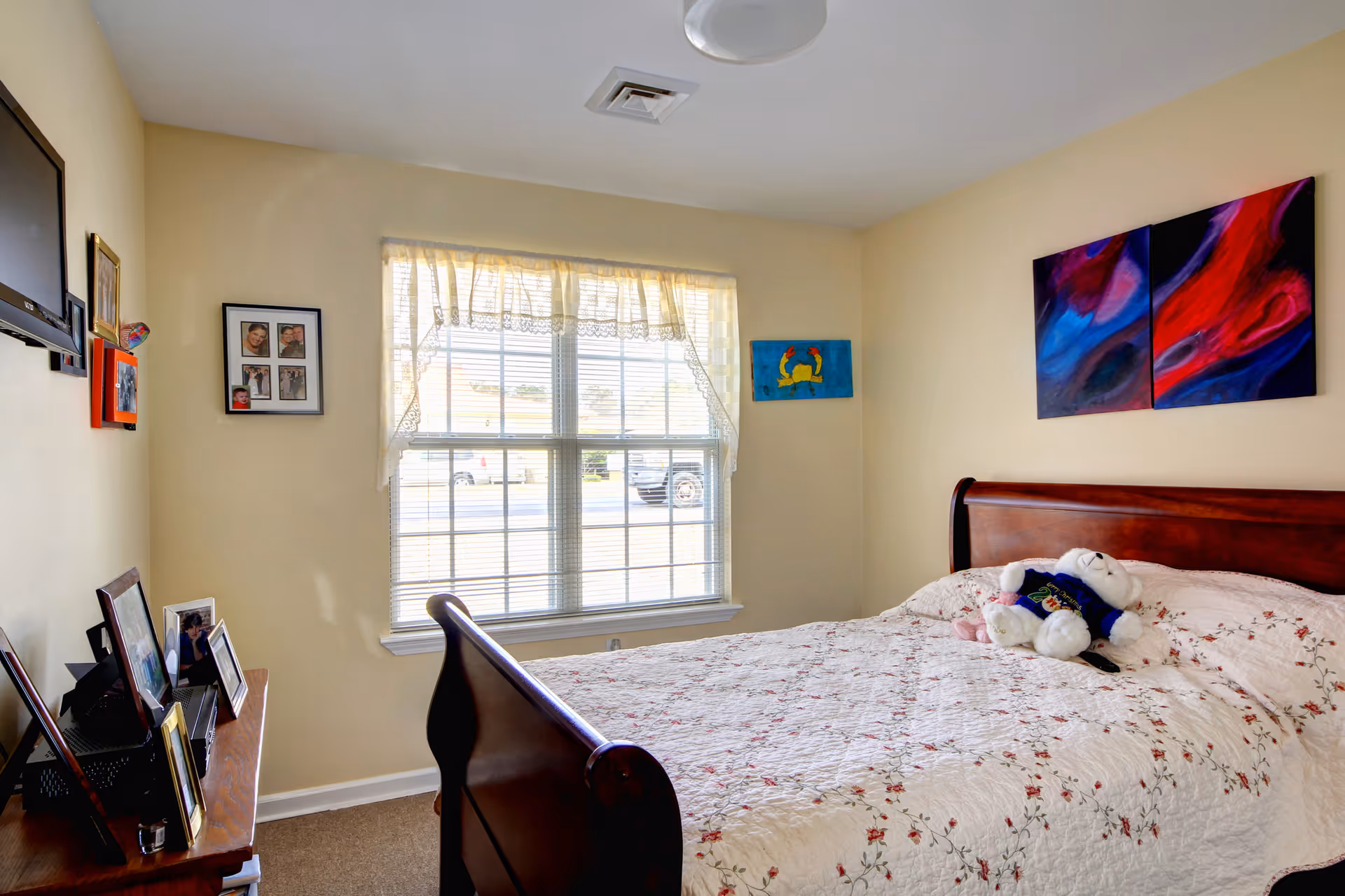 A bedroom with a wooden sleigh bed covered with a white floral quilt and a teddy bear wearing a blue sweater sitting on the bed. There is a window with sheer curtains letting in natural light. On the wall above the bed are two colorful abstract paintings. A flat-screen TV is mounted on the opposite wall above a wooden dresser with several framed photos.
