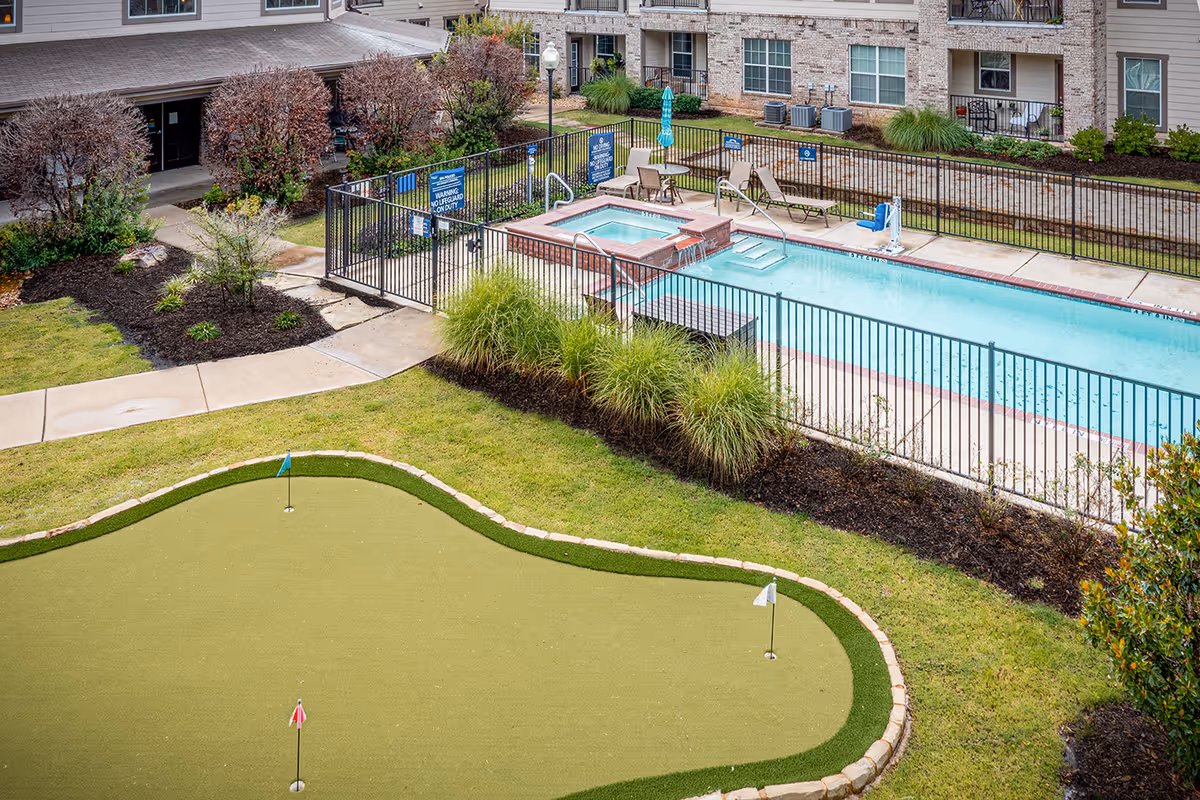 Outdoor area of Parkview on Hollybrook featuring a fenced swimming pool with lounge chairs and an adjacent hot tub. In the foreground, there is a putting green with three holes marked by small flags. Surrounding the area are landscaped bushes, grass, and a multi-story building with windows and balconies.