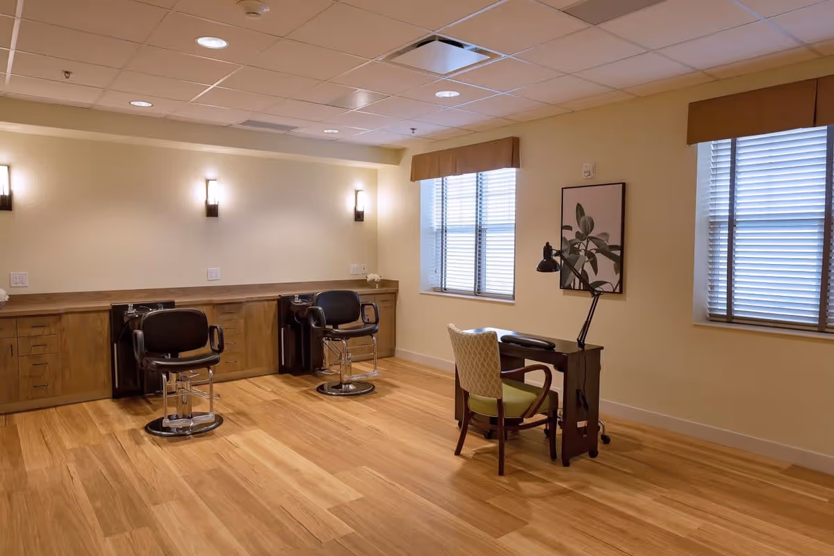 A well-lit room with wooden flooring featuring two black salon chairs in front of a long wooden counter with sinks, and a small desk with a green cushioned chair and a desk lamp near two windows with blinds.