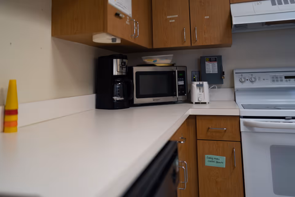 A kitchen countertop with a coffee maker, microwave, and toaster. Wooden cabinets are mounted above and below the counter. A white stove and oven are visible on the right side. A small note is attached to one of the lower cabinet doors.
