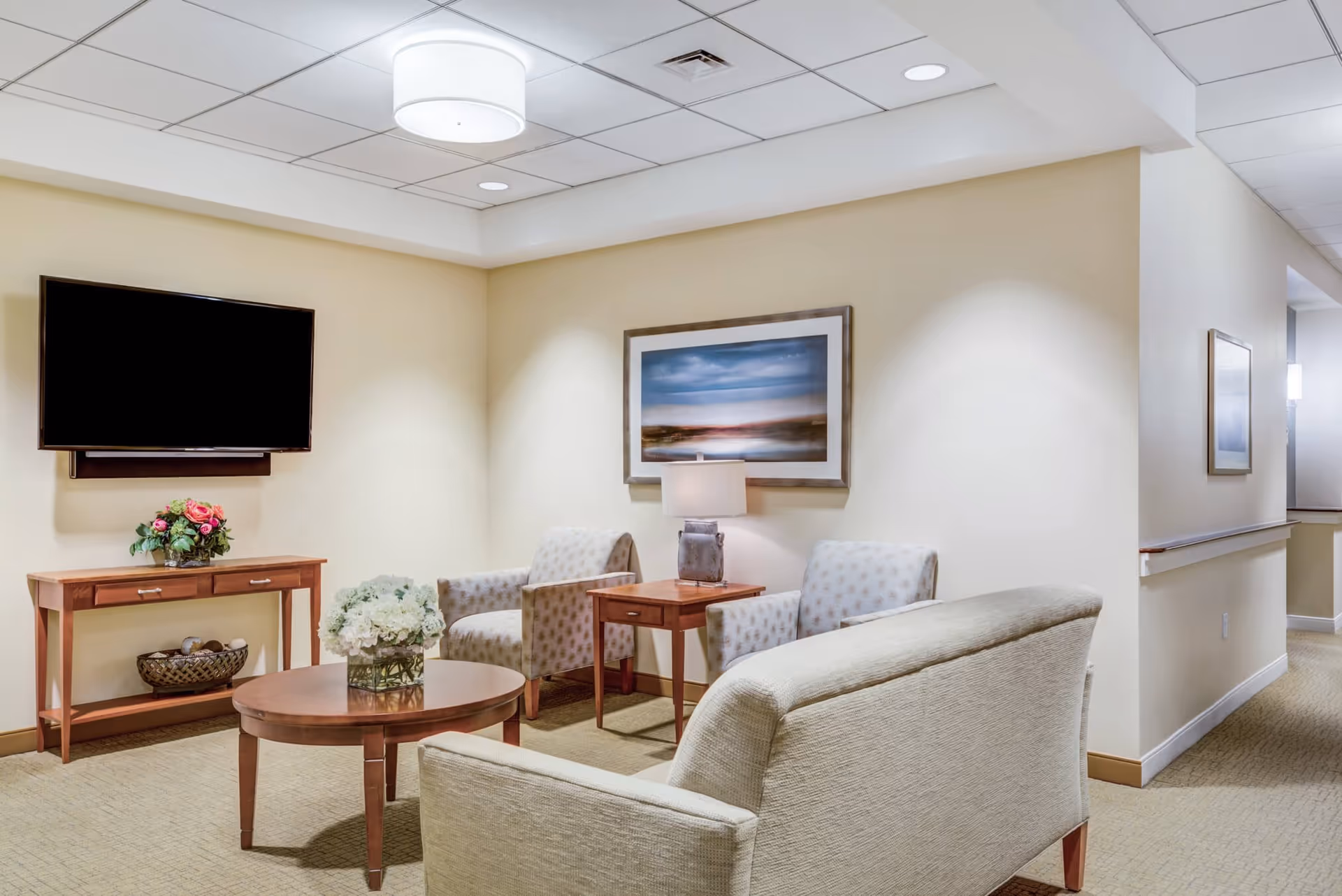 A cozy senior living common area with a beige sofa, two patterned armchairs, a wooden coffee table with a flower arrangement, a side table with a lamp, a wall-mounted flat-screen TV, and framed artwork on the walls. The room has soft lighting and neutral-colored walls and carpet.