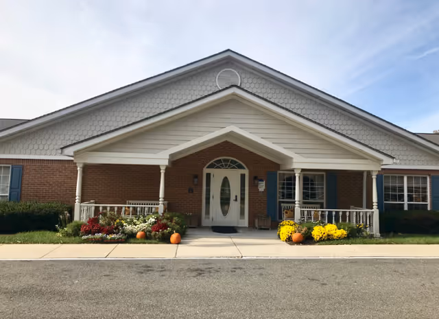 Front entrance of a single-story brick building with a covered porch, white columns, pumpkins and yellow flowers.