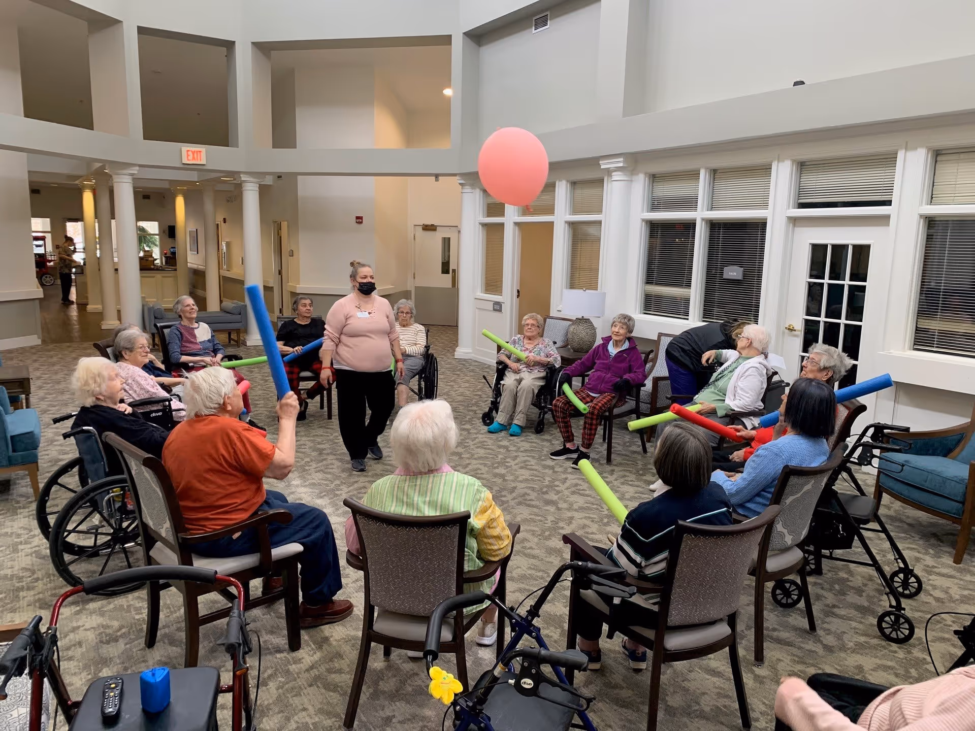 A group of elderly people seated in a circle in a spacious, well-lit common area, participating in a group activity with colorful foam noodles and a large pink balloon. A staff member wearing a mask stands in the middle, facilitating the activity.