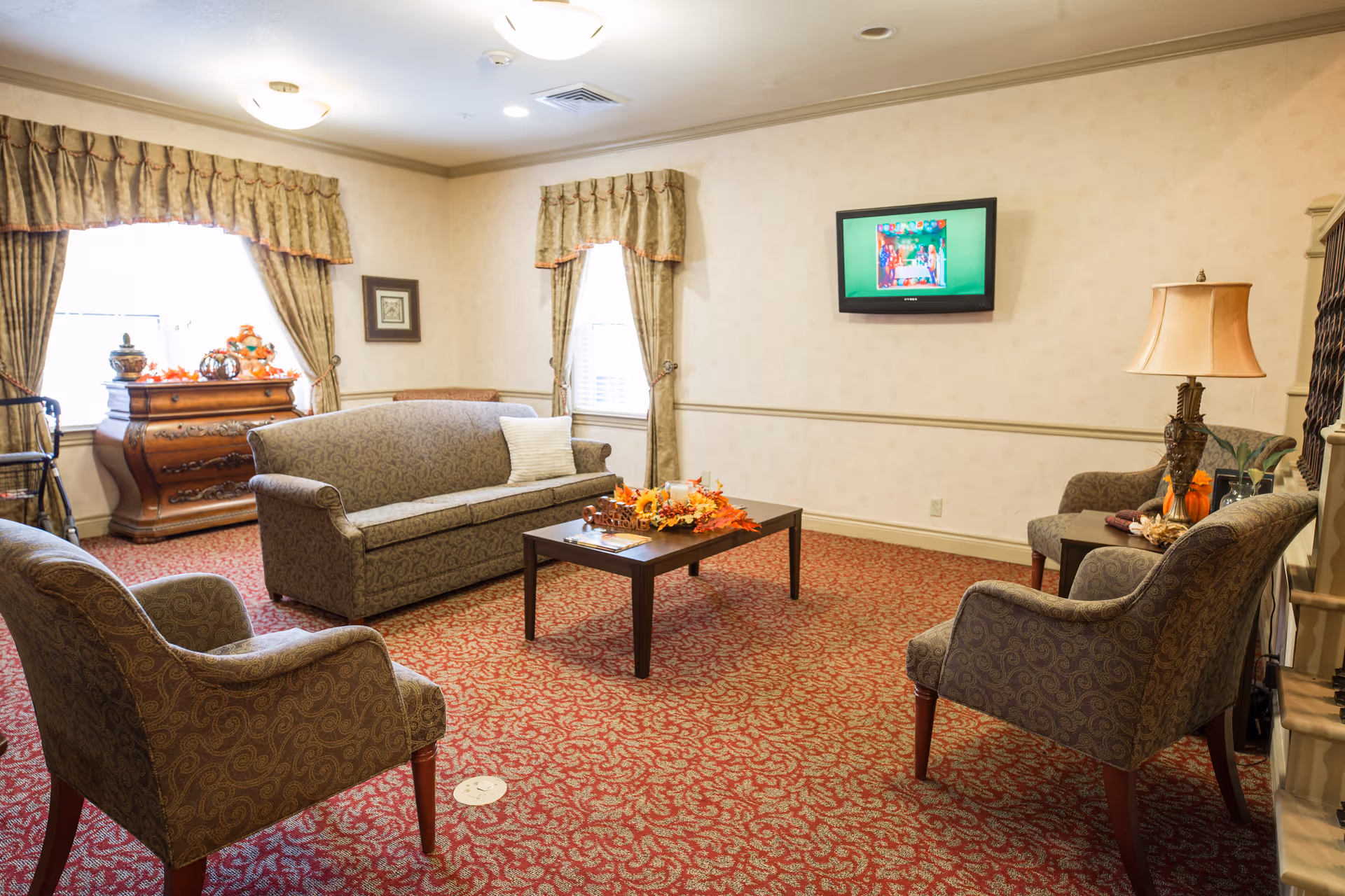 A cozy living room with patterned carpet and wallpaper. The room features a sofa, two armchairs, a wooden coffee table decorated with autumn-themed items, a side table with a lamp, and a wooden chest of drawers near two windows with curtains. A flat-screen TV is mounted on the wall.