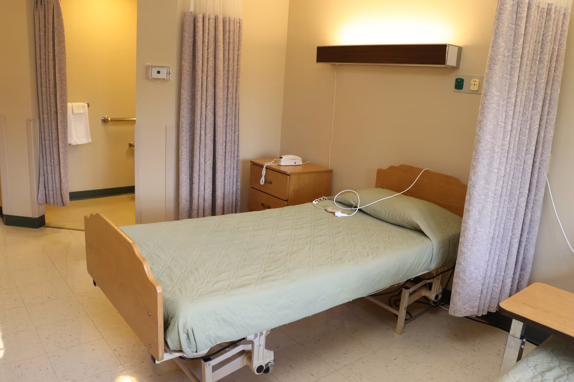 A simple medical-style single bed with green bedding in a private patient room with a bedside table, privacy curtains, and an adjacent bathroom entrance.