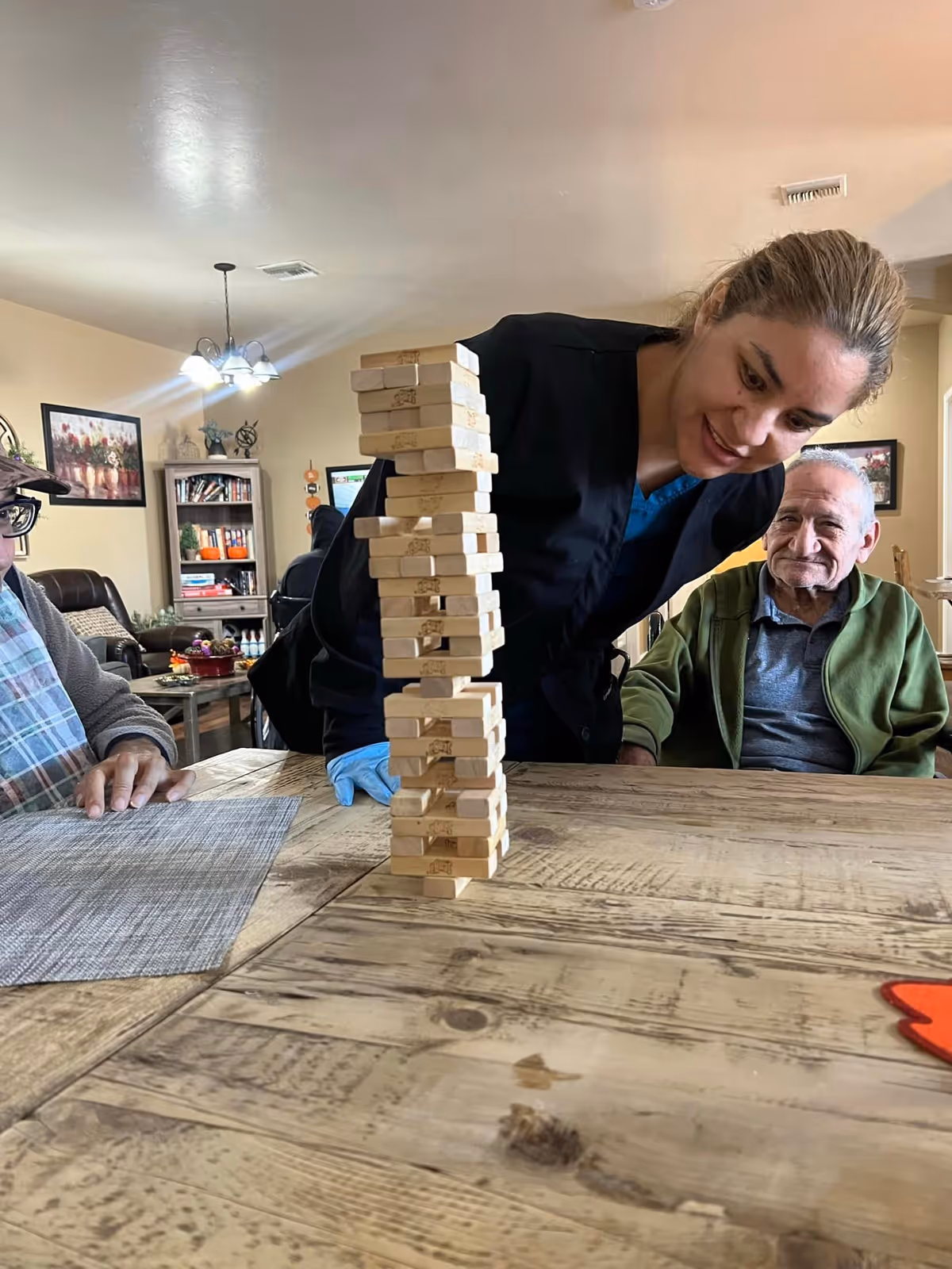 A woman in a black uniform and blue gloves is leaning over a wooden table, carefully playing a game of Jenga with two elderly men seated at the table in a cozy living room setting. The room has warm lighting, a bookshelf, framed pictures on the walls, and a chandelier overhead.