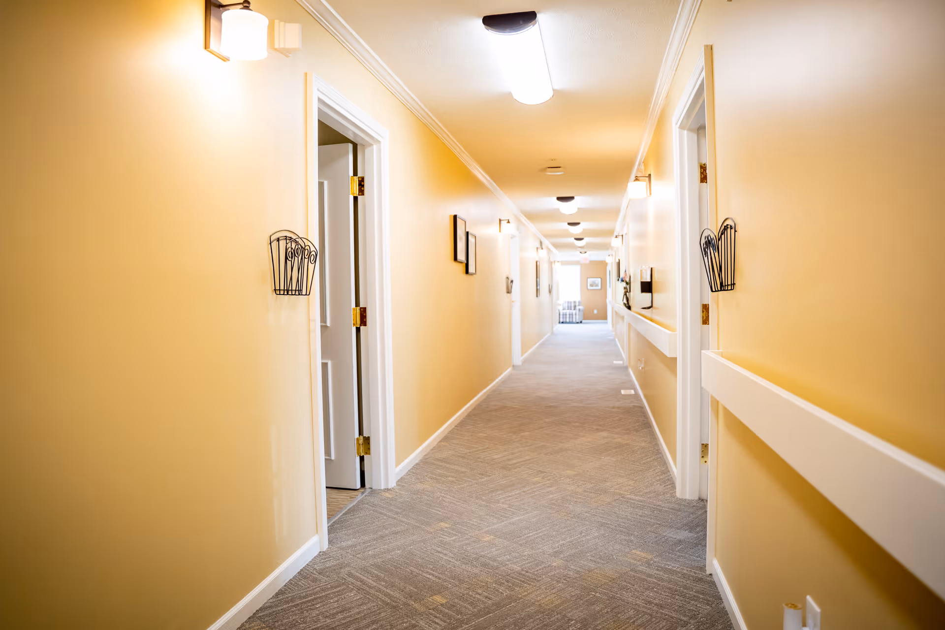 Well-lit yellow-painted interior hallway with open doors and carpeted floor leading to a distant seating area.