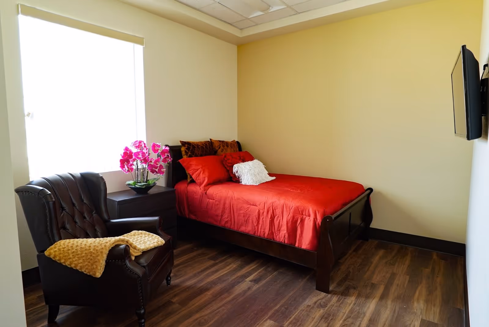 A small bedroom with a single bed covered in a red bedspread and multiple pillows, a dark wooden nightstand with a pink orchid plant, a dark brown leather armchair with a yellow blanket draped over one arm, and a wall-mounted flat screen TV. The room has light yellow walls and wood flooring.