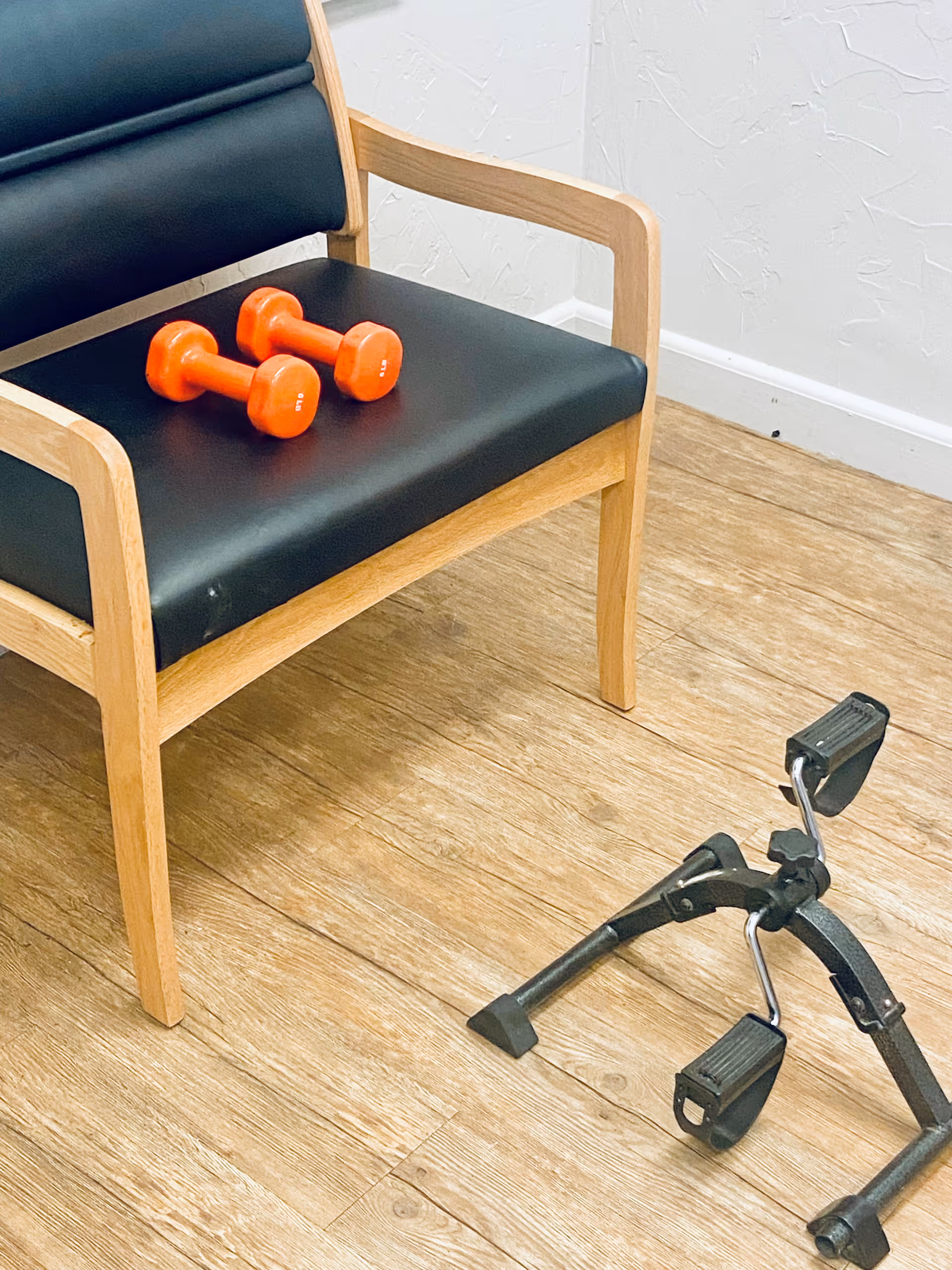 A wooden chair with black cushioned seat and backrest, with two small orange dumbbells placed on the seat. On the wooden floor next to the chair is a compact pedal exerciser.
