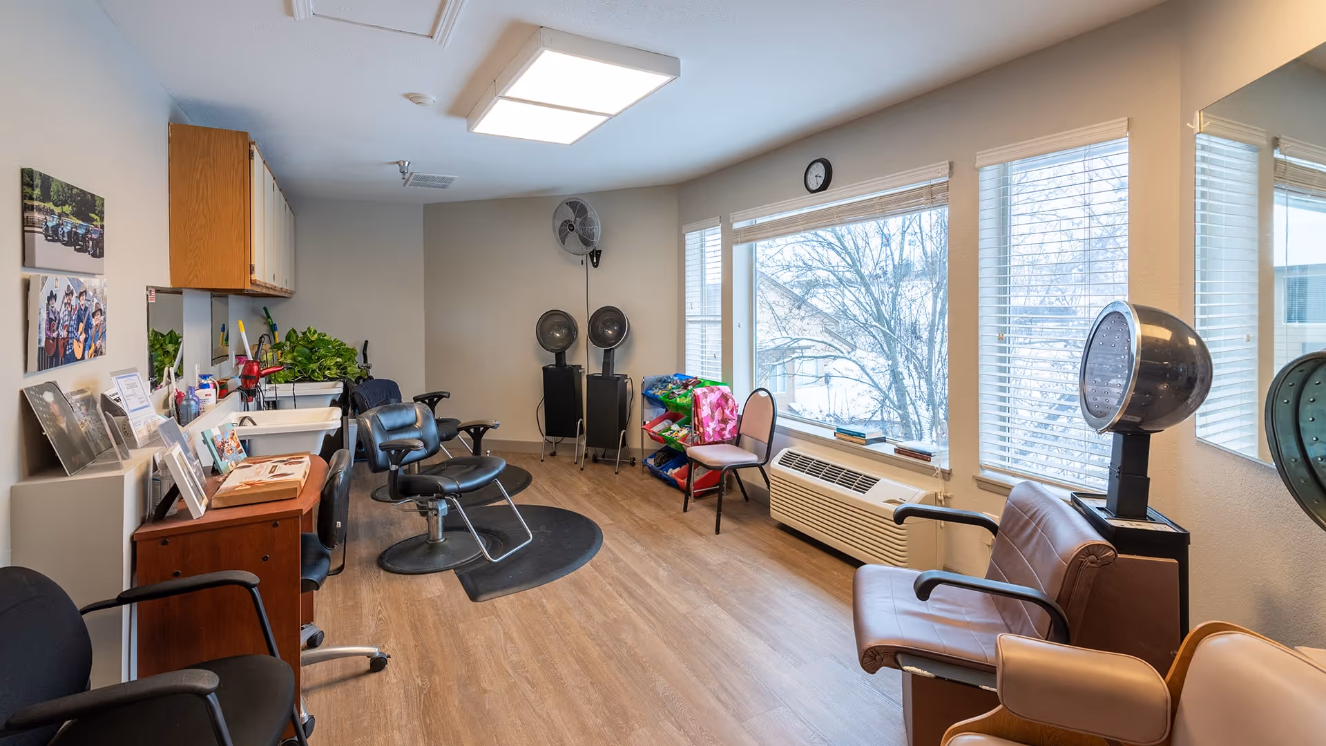 Interior view of a hair salon area in a senior living facility with salon chairs, hair dryers, a wooden desk with framed photos, plants, and large windows with blinds letting in natural light.