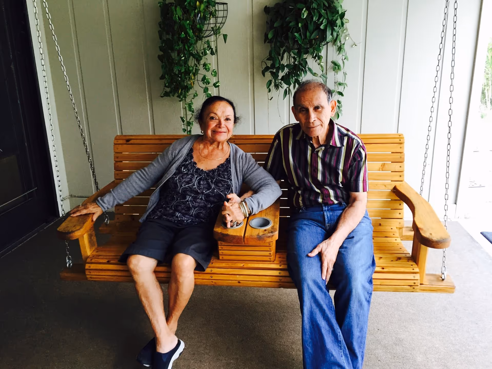 An elderly woman and man sitting together on a wooden porch swing with hanging plants in the background. The woman is smiling and holding the man's hand, and they are seated on a covered porch area.
