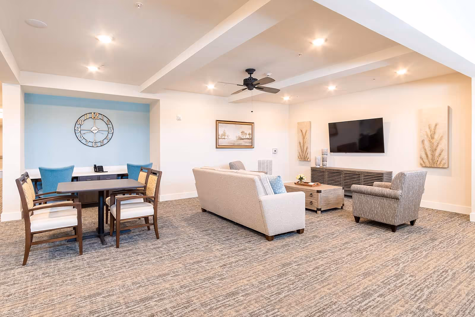 A bright and spacious living room area with a beige sofa, a gray armchair, a wooden coffee table, and a wall-mounted flat screen TV. The room has a ceiling fan, recessed lighting, and light-colored walls with decorative artwork. To the left, there is a small dining or game table with four chairs, two of which are blue and two with wooden frames and beige cushions. A large decorative clock is mounted on a light blue accent wall behind the table.