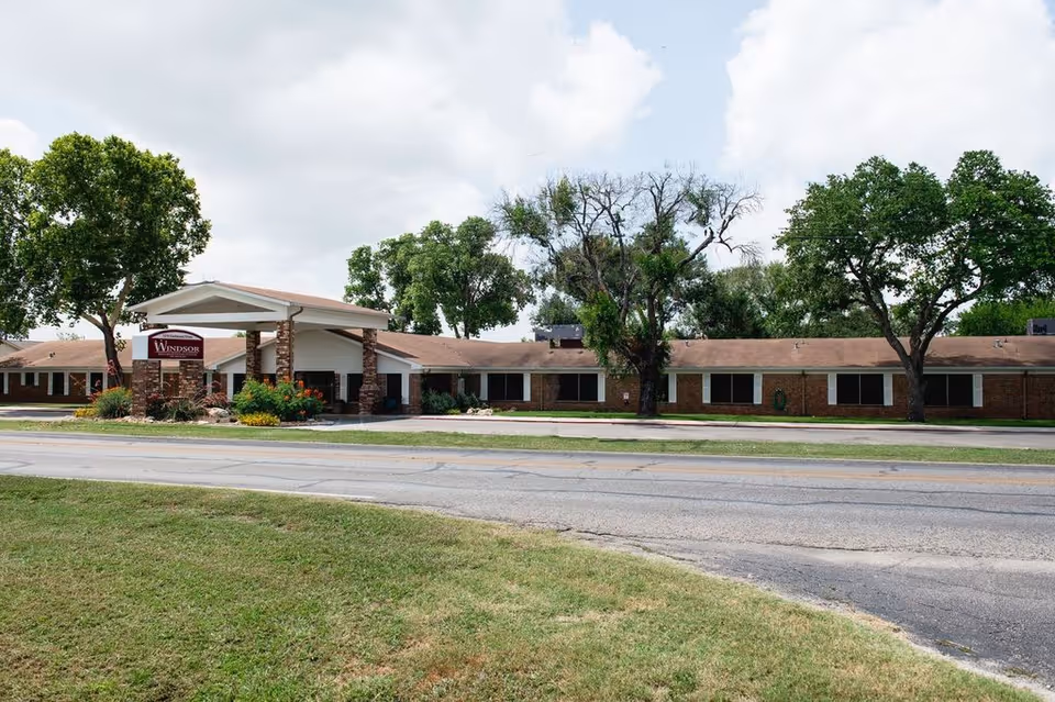 Exterior view of Windsor Nursing and Rehabilitation Center of Seguin, a single-story brick building with a covered entrance supported by stone pillars, surrounded by green trees and grass under a partly cloudy sky.