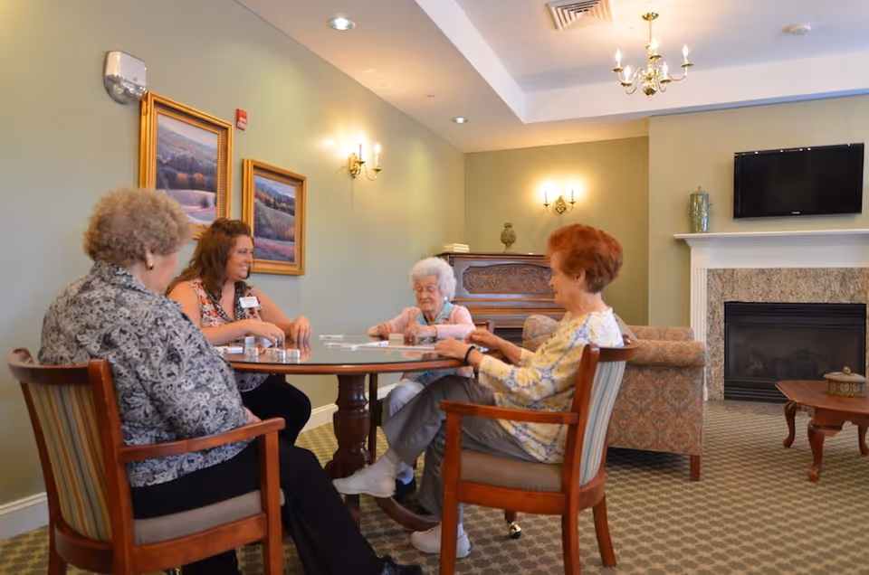 Four elderly women sitting around a round wooden table playing a game in a cozy living room with green walls, framed landscape paintings, a fireplace, and a mounted TV.