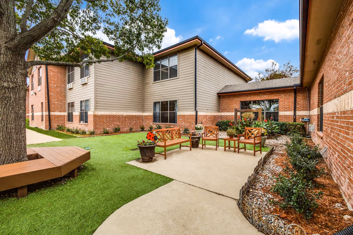 Outdoor courtyard area of a senior living facility with a large tree surrounded by a wooden bench, several wooden chairs and benches arranged on a concrete patio, potted plants, and well-maintained landscaping including grass and shrubs. The building exterior features brick and siding with multiple windows under a partly cloudy blue sky.