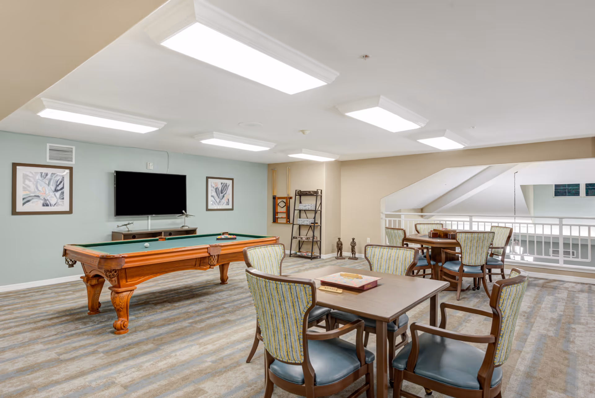 A recreational room with a pool table in the center, a large flat-screen TV mounted on a light blue wall, and two framed artworks on either side. There are two tables with chairs around them, one with a Scrabble game set up. The room has bright overhead lighting and a carpeted floor with a striped pattern. A railing overlooks a lower level in the background.
