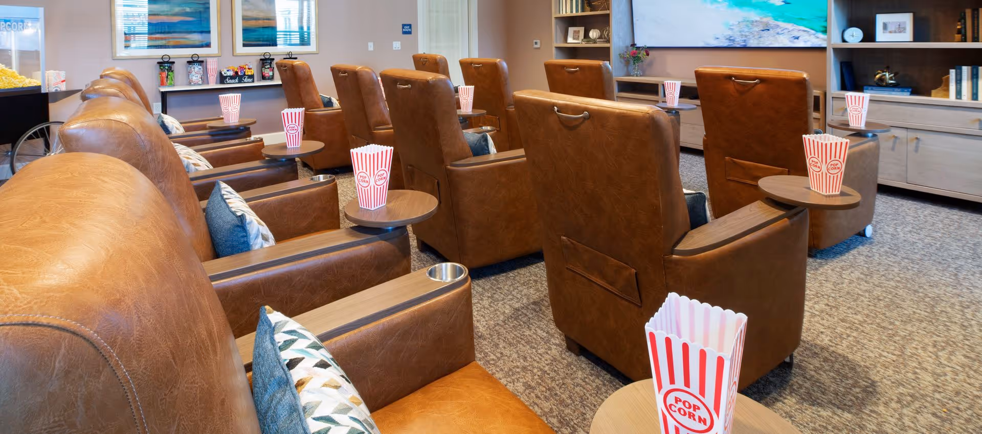 A cozy movie theater room with multiple rows of brown leather recliner chairs, each equipped with a small wooden side table holding a popcorn container. The room features a large flat-screen TV on the wall, shelves with books and decorative items, and a popcorn machine in the corner.