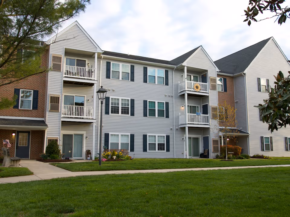 Exterior view of a three-story residential building with balconies, multiple windows, and a well-maintained lawn in front. There is a sidewalk, a street lamp, and some trees and shrubs around the building.