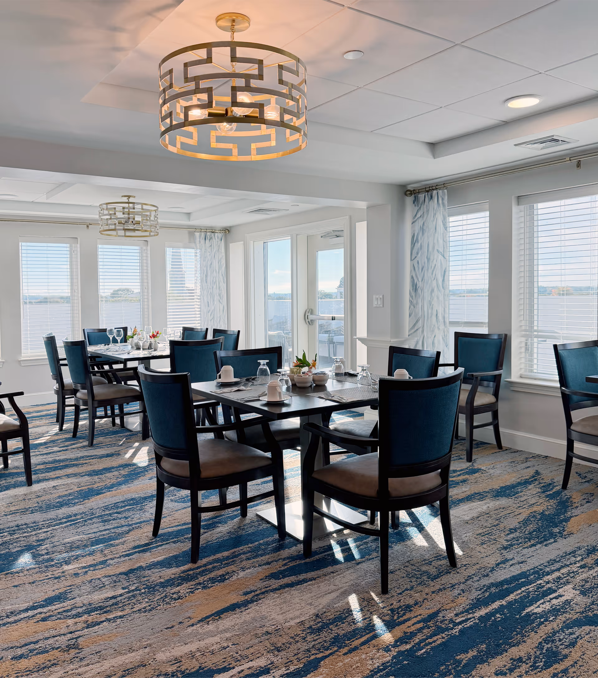 Bright dining room with multiple tables and blue-upholstered chairs, large windows and modern chandeliers overlooking the water.