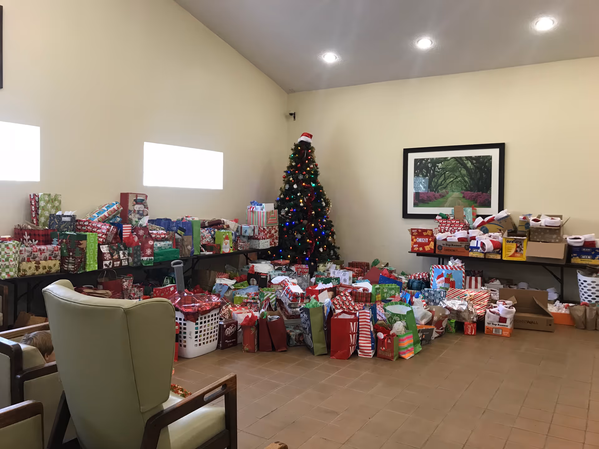 Community room with a decorated Christmas tree surrounded by numerous wrapped gifts and chairs.