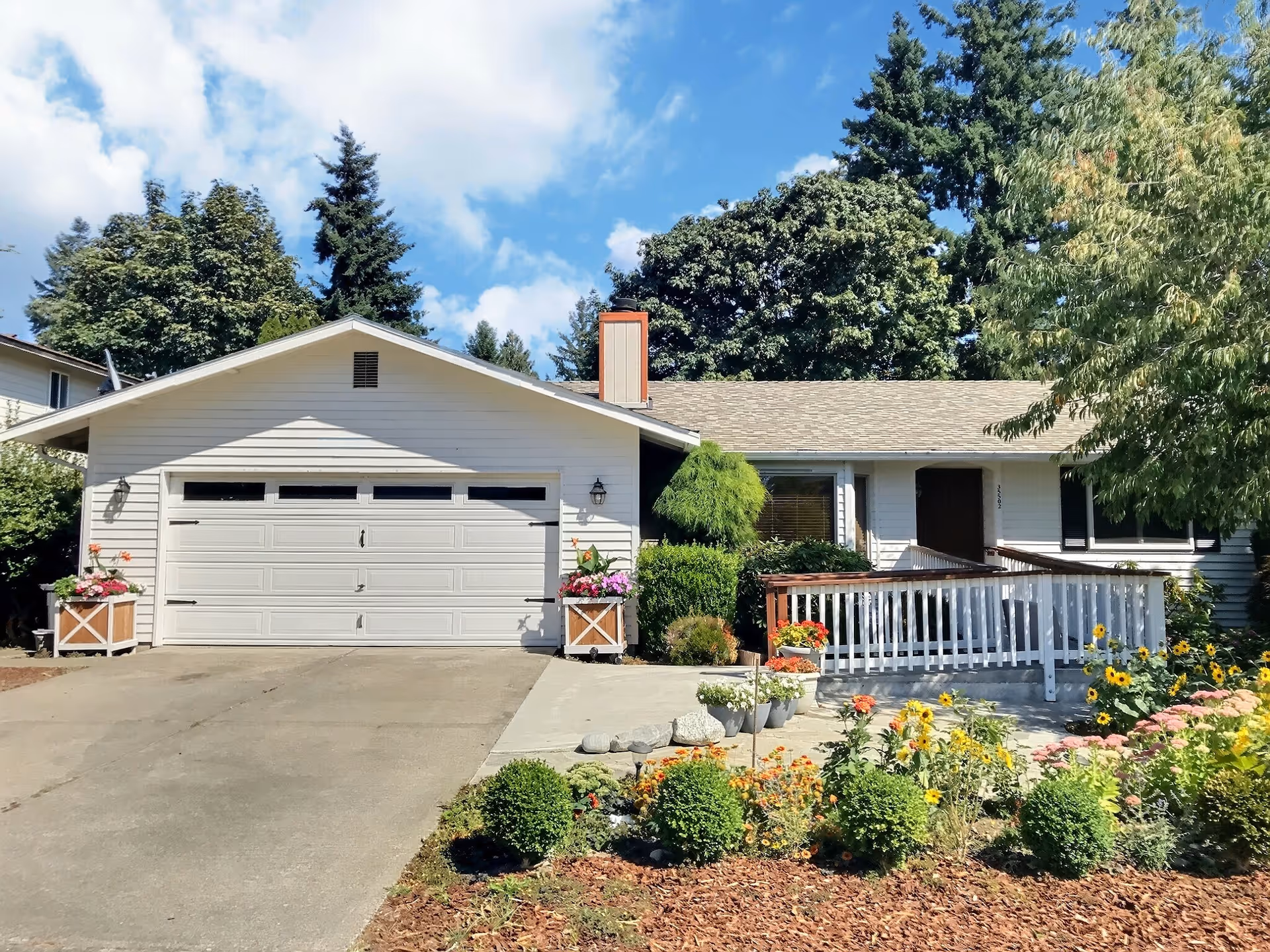 Front exterior view of a single-story house with a two-car garage, a ramp leading to the front door, and a well-maintained garden with various flowers and shrubs under a partly cloudy sky.