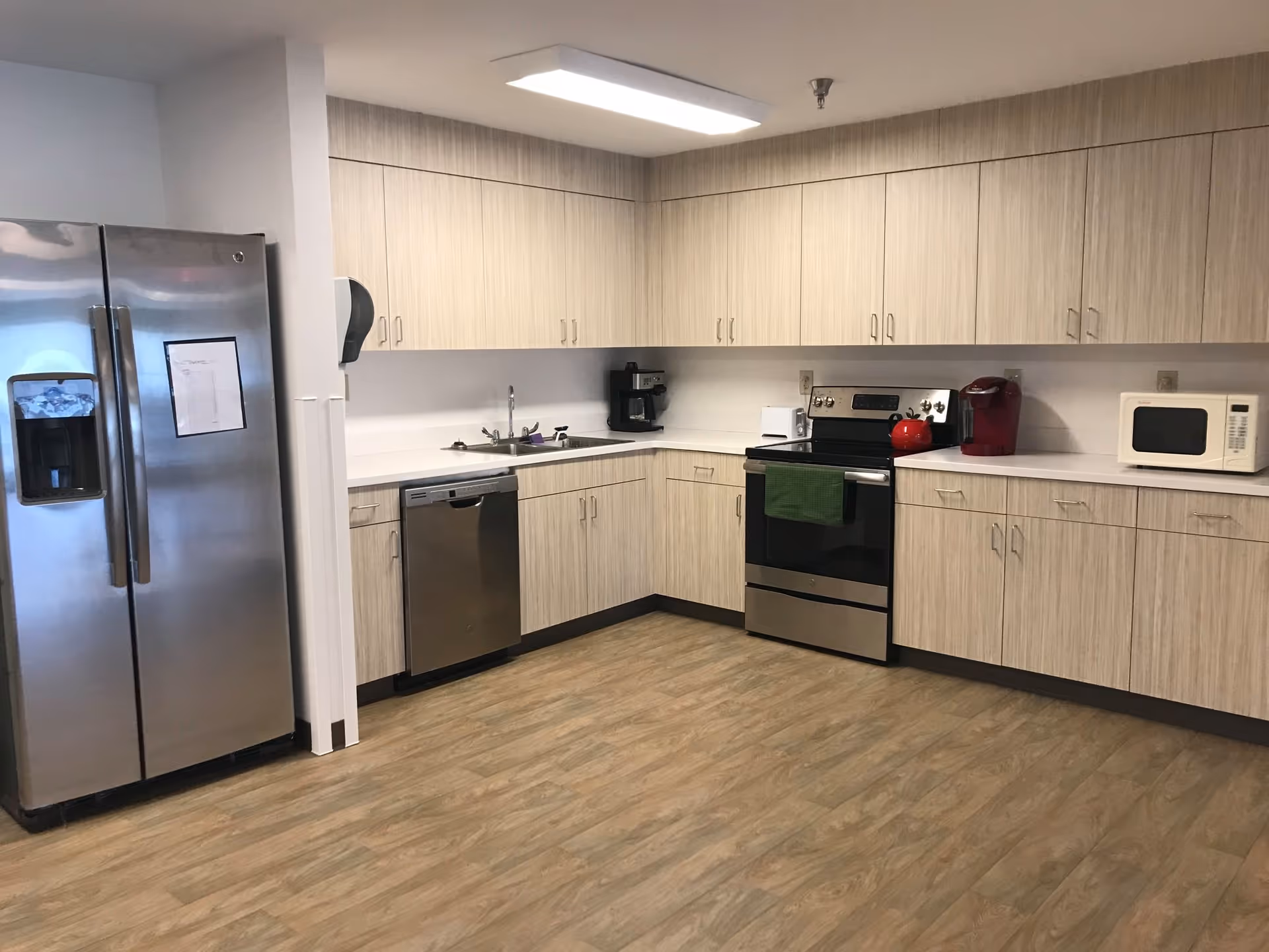 A modern kitchen with light wood cabinets, stainless steel refrigerator, dishwasher, and oven. The kitchen also features a microwave, coffee maker, and a red kettle on the stove. The floor is wood-patterned, and the walls are light-colored.