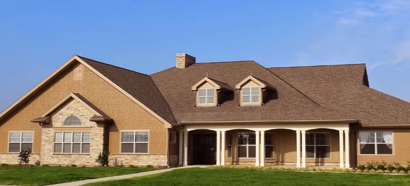Front exterior of a single-story tan building with a covered porch, columns, dormer windows, and a green lawn under a blue sky.