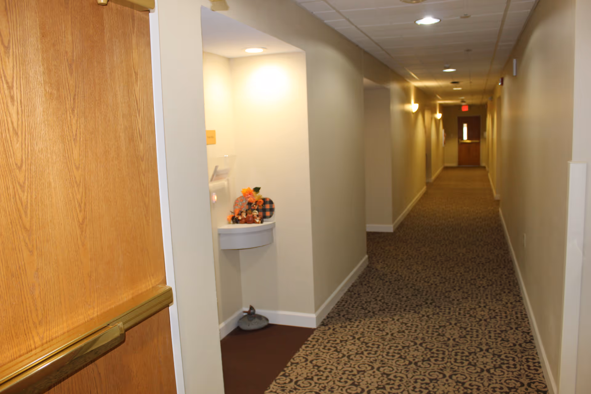 A long, carpeted hallway in a senior living facility with beige walls and ceiling lights. On the left side near the foreground, there is a small wall-mounted shelf with autumn-themed decorations including a small pumpkin and flowers. The hallway extends to a wooden door at the far end.
