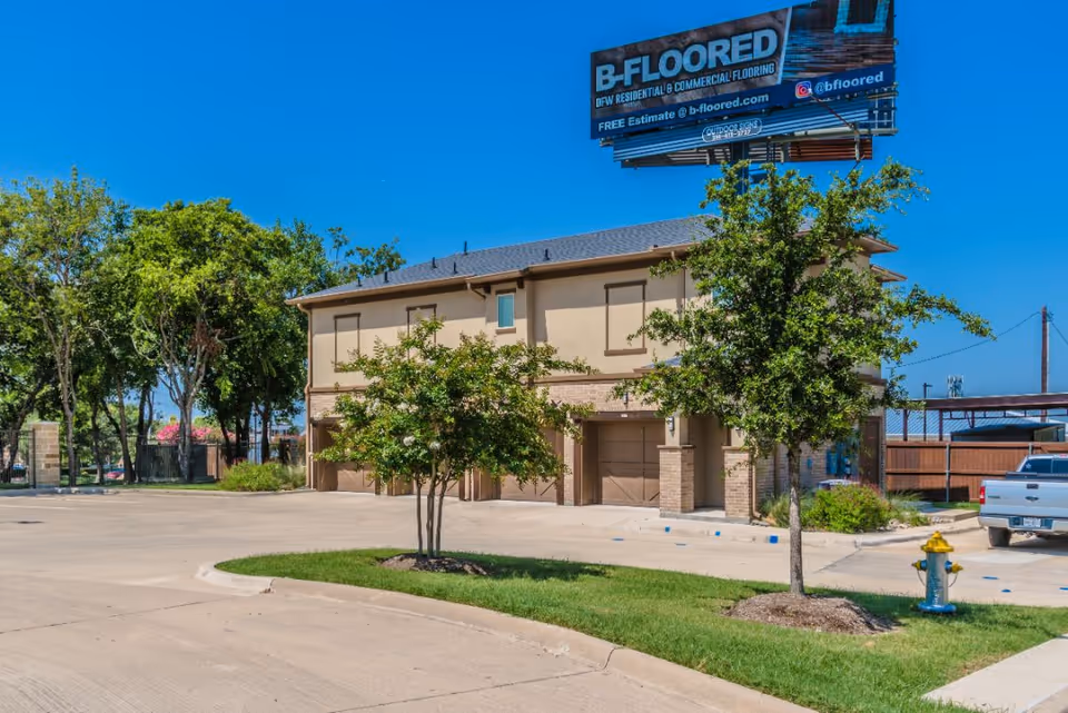 Exterior view of a two-story building with beige walls and brown trim, surrounded by trees and a parking area with a fire hydrant and a parked white pickup truck. A large billboard advertising B-FLOORED residential and commercial flooring is visible above the building against a clear blue sky.
