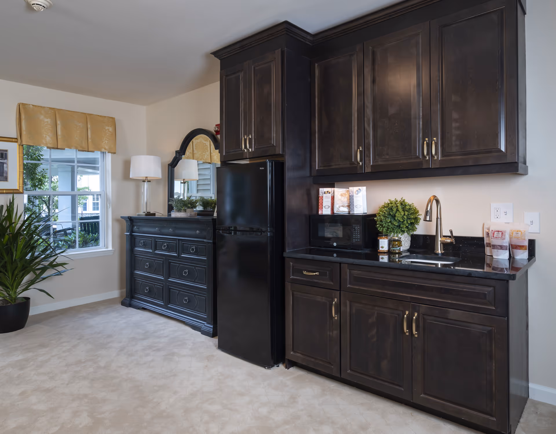 Interior view of a room featuring dark wood cabinetry with a black refrigerator, a microwave, a small sink with a faucet, and various packaged food items on the countertop. To the left, there is a black dresser with a large mirror and a table lamp, next to a window with a yellow valance and a potted plant on the floor.