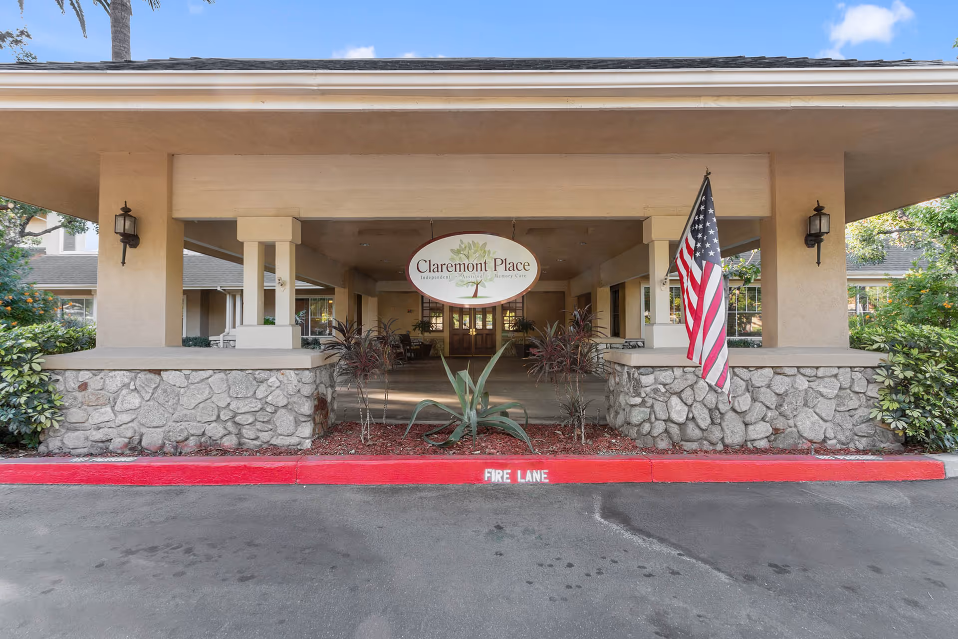 Covered front entrance of Claremont Place featuring an oval sign, an American flag, stone planters and a red fire lane curb.