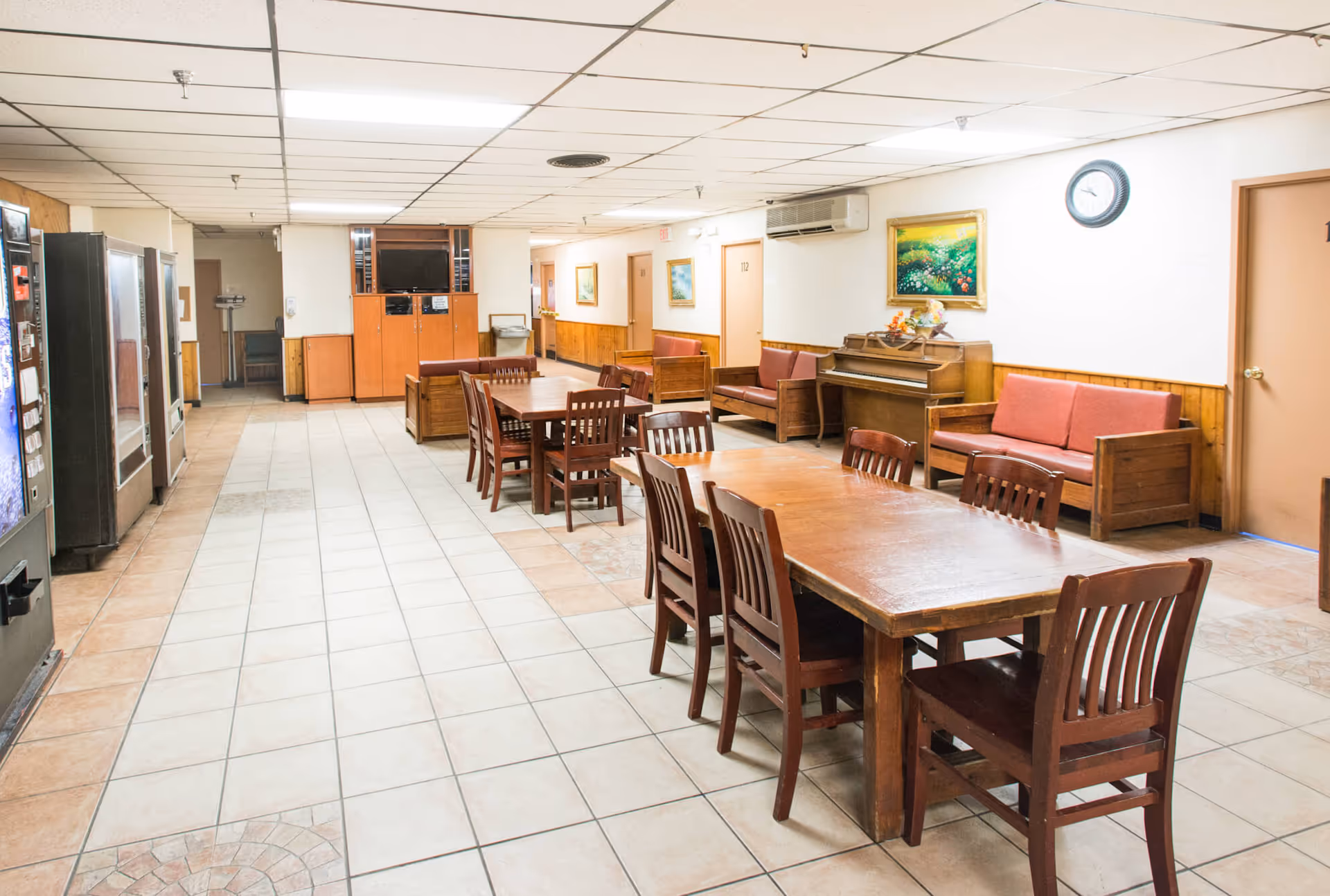 Interior of a senior living facility common area with wooden tables and chairs arranged in the center. Along the right wall, there are wooden sofas with red cushions, a piano, a wall clock, and framed paintings. The floor is tiled, and vending machines are visible on the left side. The ceiling has fluorescent lighting and a white grid pattern.