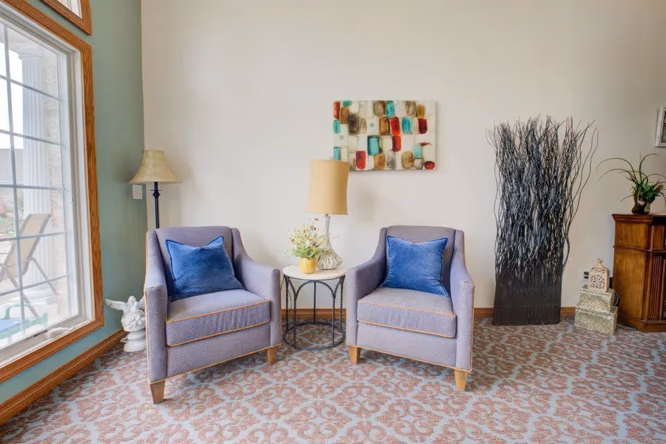 A cozy seating area with two lavender armchairs each with a blue cushion, separated by a small round table holding a lamp and a small flower arrangement. The area features a patterned carpet, a floor lamp, a decorative wall art piece, and a tall vase with decorative branches. A large window on the left side lets in natural light.