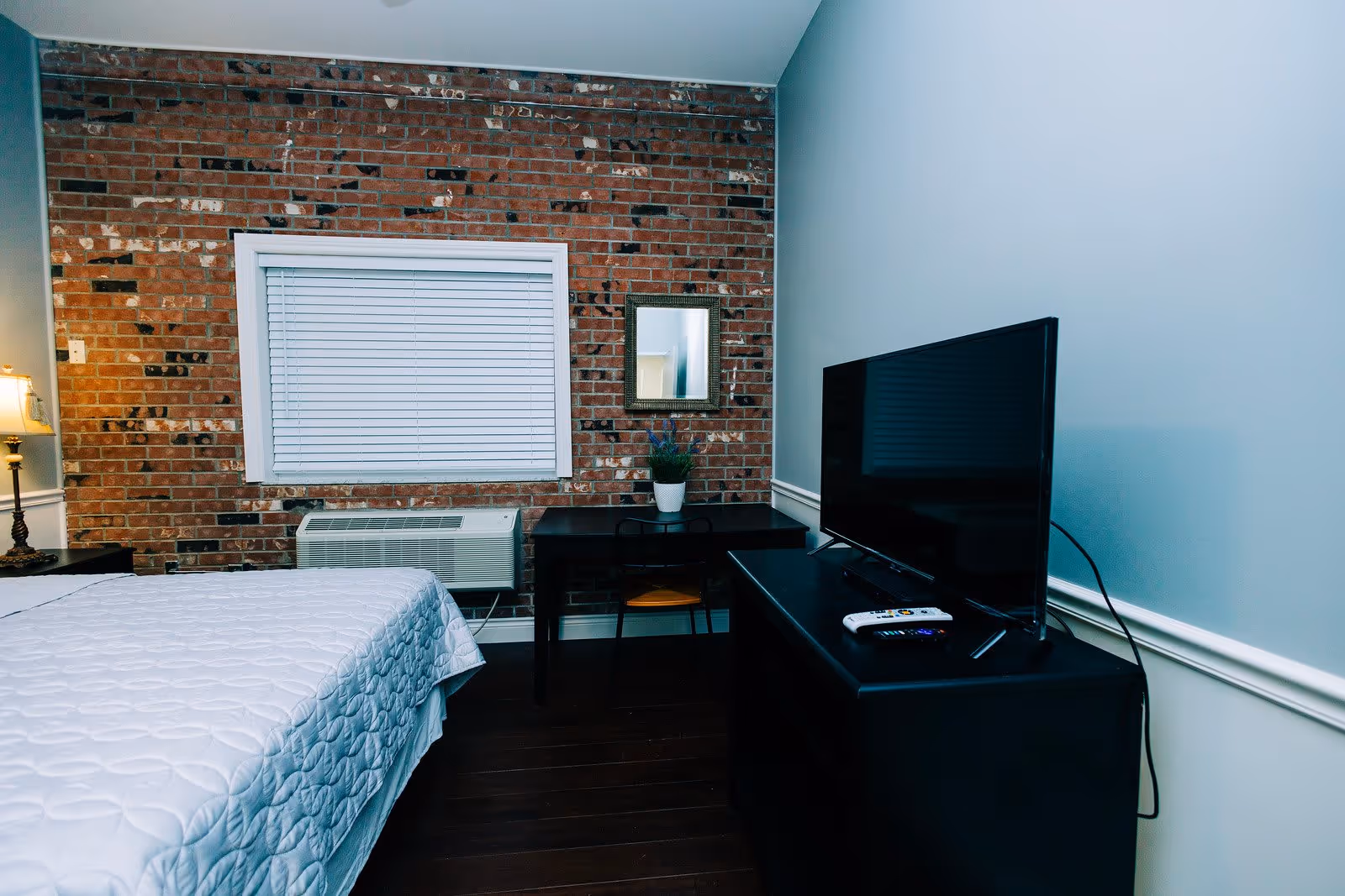 A bedroom with a bed covered in a white quilted bedspread, a brick accent wall with a window and white blinds, a black desk with a chair and a small potted plant, a mirror on the wall, and a black dresser with a flat-screen TV and remote controls on top.