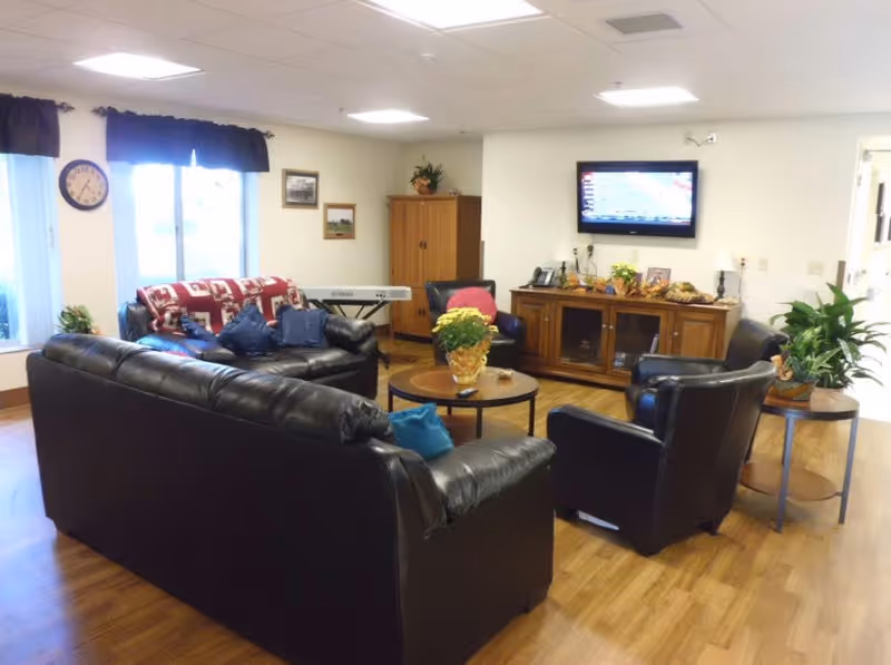 A cozy living room area with black leather sofas and chairs arranged around a round wooden coffee table with a potted plant. A wooden cabinet with a flat-screen TV mounted above it is against the wall. There are windows with dark curtains, a clock on the wall, and a keyboard in the corner. The floor is wooden, and there are additional plants and framed pictures on the walls.