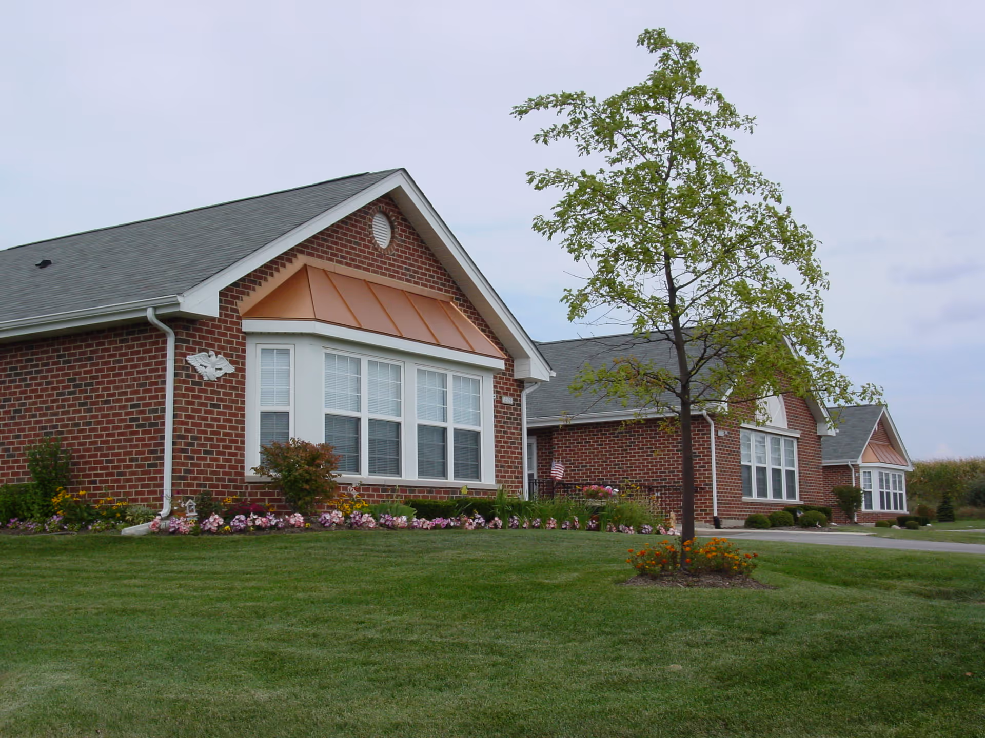 Exterior view of a brick building with white-trimmed windows and a gray roof, surrounded by a well-maintained lawn, flower beds, and a young tree in the foreground under a cloudy sky.