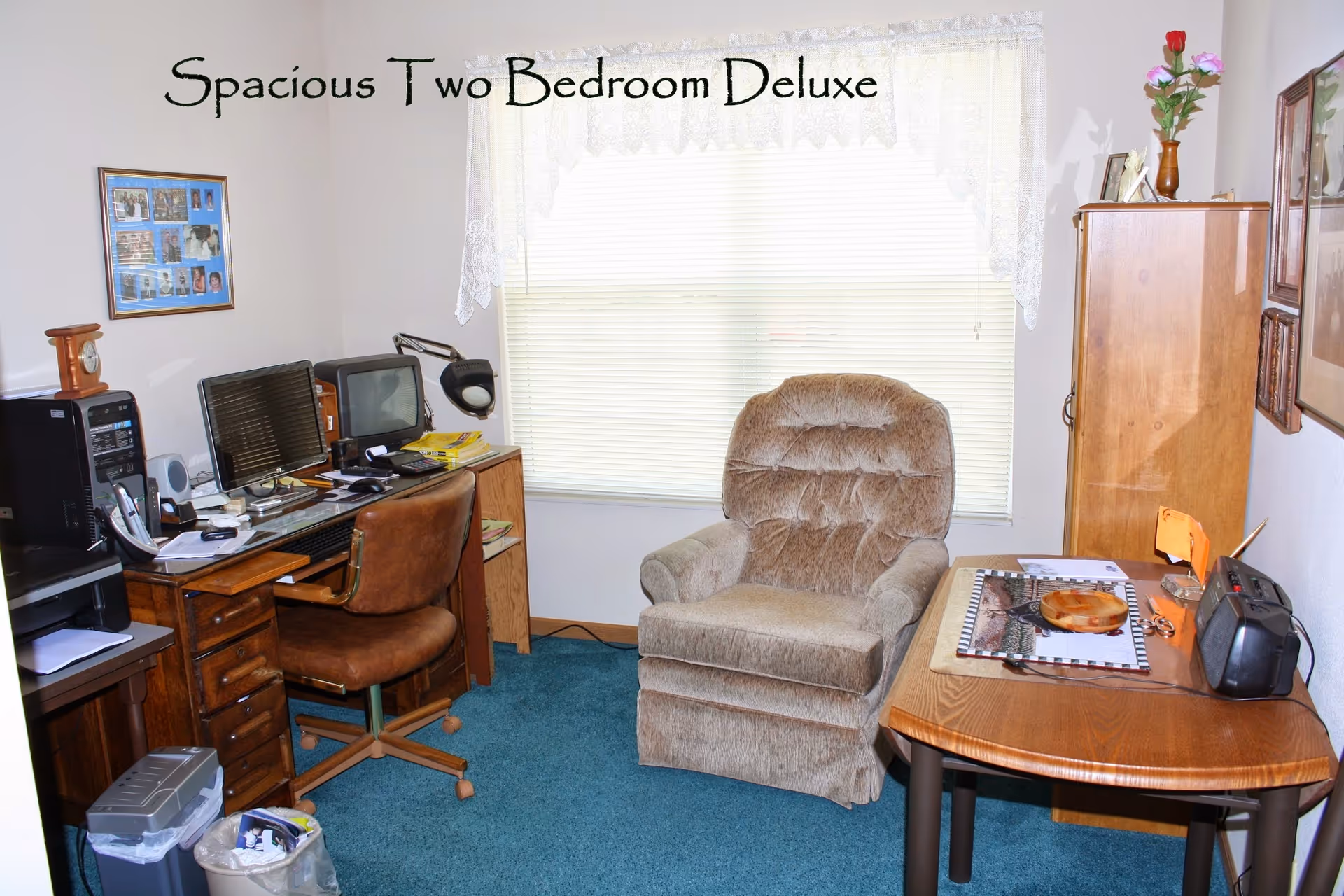 A cozy room with a brown cushioned armchair in front of a window with white blinds and lace curtains. To the left is a wooden desk with a brown swivel chair, computer monitors, and various office supplies. On the right side, there is a wooden table with a radio, papers, and a small wooden cabinet with a vase holding flowers on top. The carpet is blue, and the walls are decorated with framed pictures. Text above the window reads 'Spacious Two Bedroom Deluxe'.