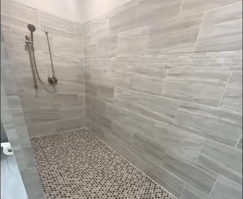 A walk-in shower with light gray rectangular wall tiles and small hexagonal floor tiles in shades of gray and brown. The shower features a handheld showerhead attached to a vertical grab bar on the left wall.