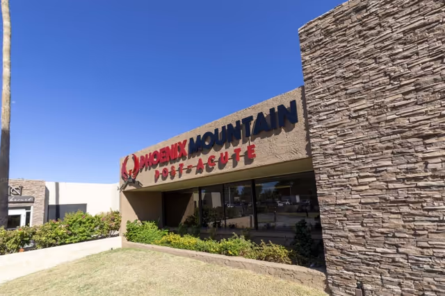 Exterior view of the Phoenix Mountain Post-Acute facility building with clear blue sky, stone and stucco walls, and landscaping with bushes and grass in front.