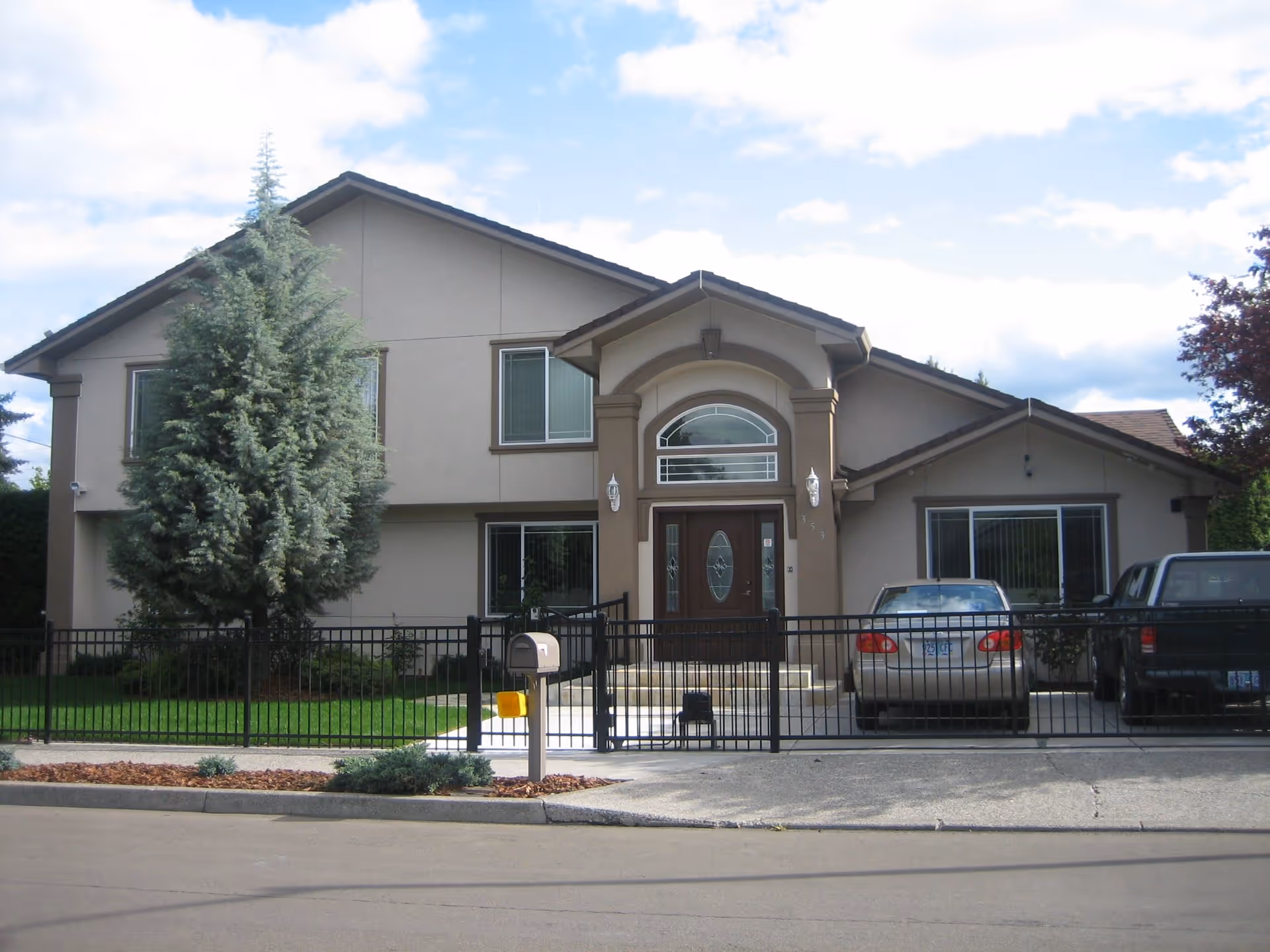 Front exterior view of a two-story residential building with a gated driveway, two parked cars, a mailbox, and a large tree in the front yard under a partly cloudy sky.