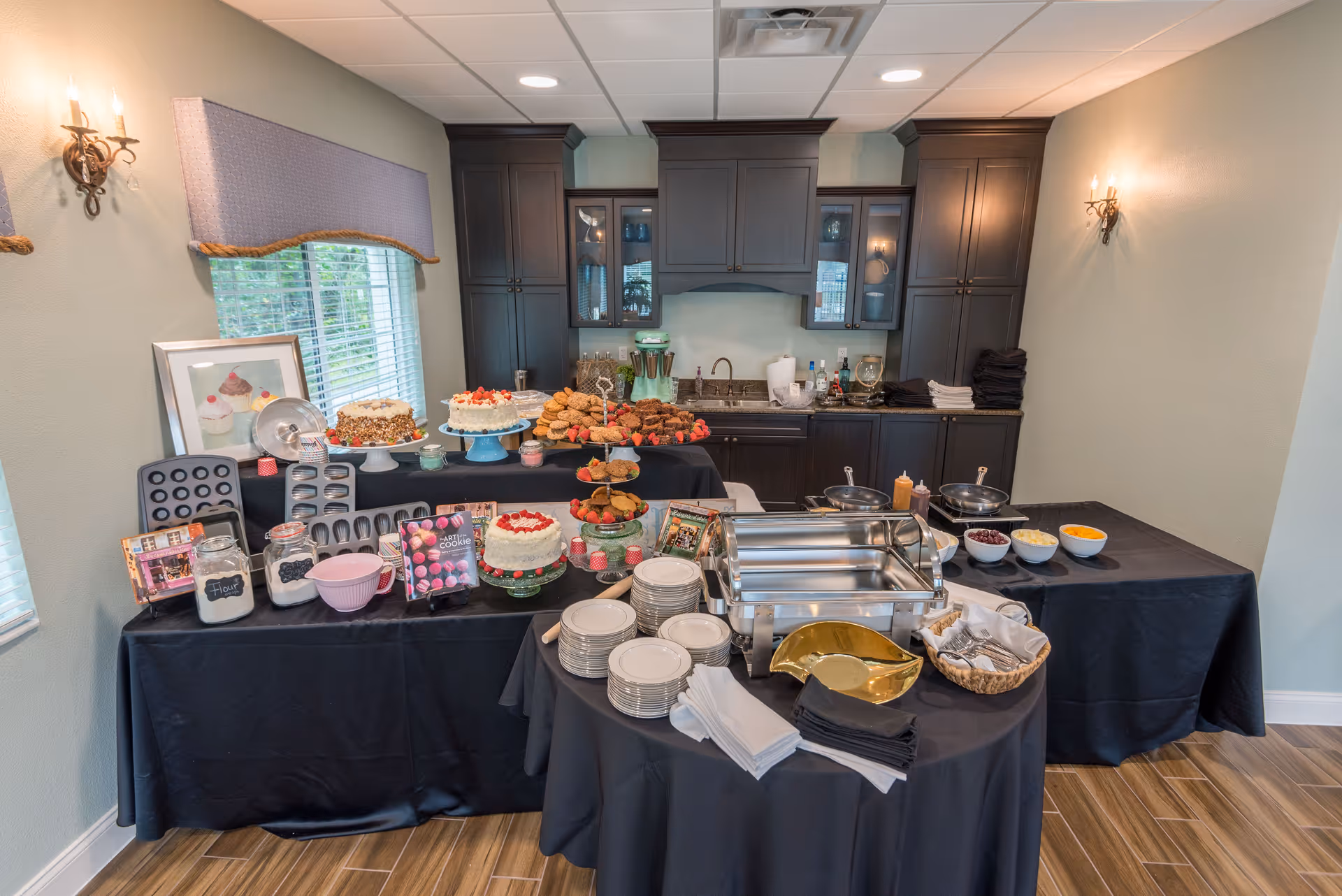 A kitchen area set up for a buffet with various desserts including cakes, cookies, and muffins displayed on tables covered with black tablecloths. There are plates, napkins, and serving utensils arranged on a round table in the foreground. The background features dark wood cabinets, a sink, and countertop with additional items.
