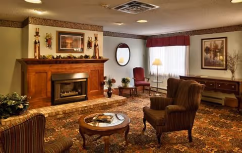Cozy senior living lounge with a fireplace, upholstered armchairs, coffee table and framed artwork.