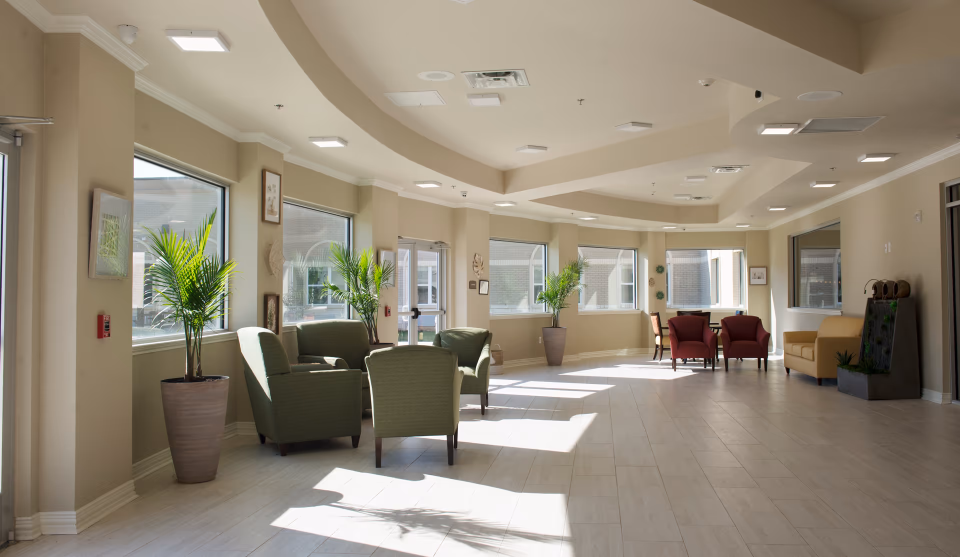 Bright, spacious senior living lounge with multiple seating areas, potted plants, and large windows.
