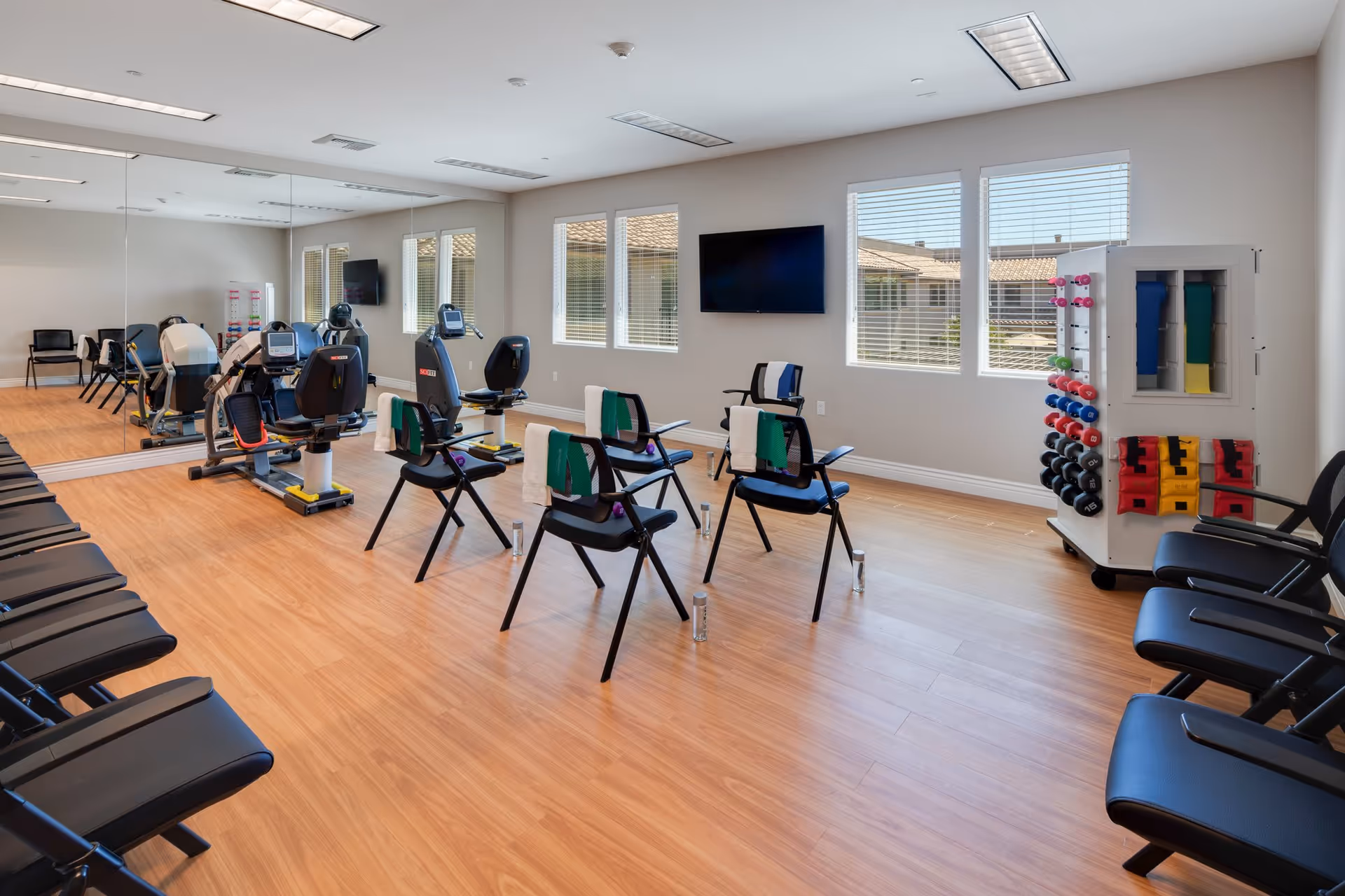 A bright exercise room with wooden flooring, several black chairs arranged in rows along the walls, and four chairs in the center equipped with green and white exercise bands. There are two recumbent exercise bikes near a large wall mirror, a TV mounted on the wall, and a rack holding colorful dumbbells and exercise mats near the windows.