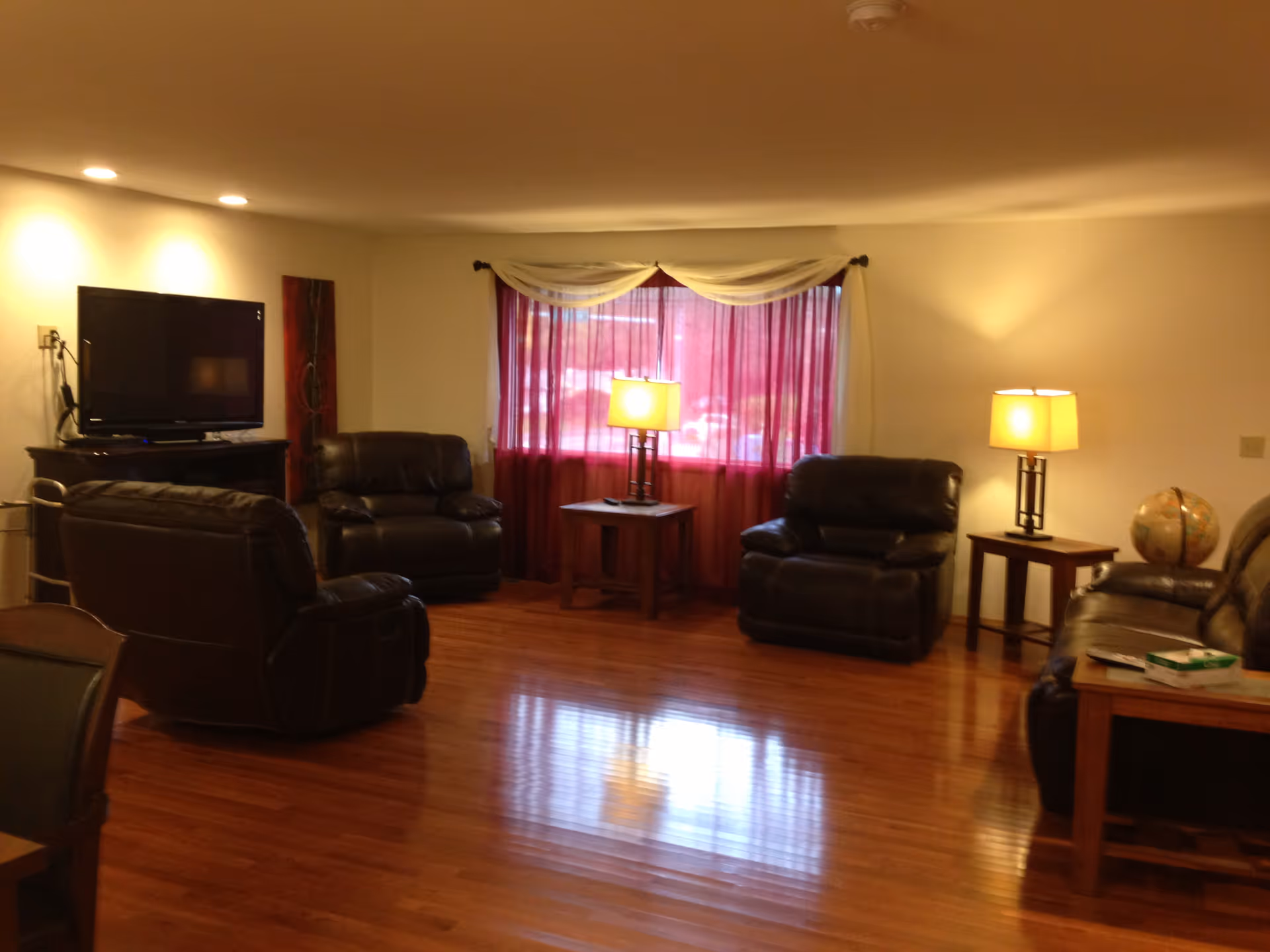 A cozy living room with polished wooden floors, three dark leather armchairs, a matching leather sofa, two wooden side tables with lamps, a globe on one table, a flat-screen TV on a wooden stand, and a window with red curtains and white valance.
