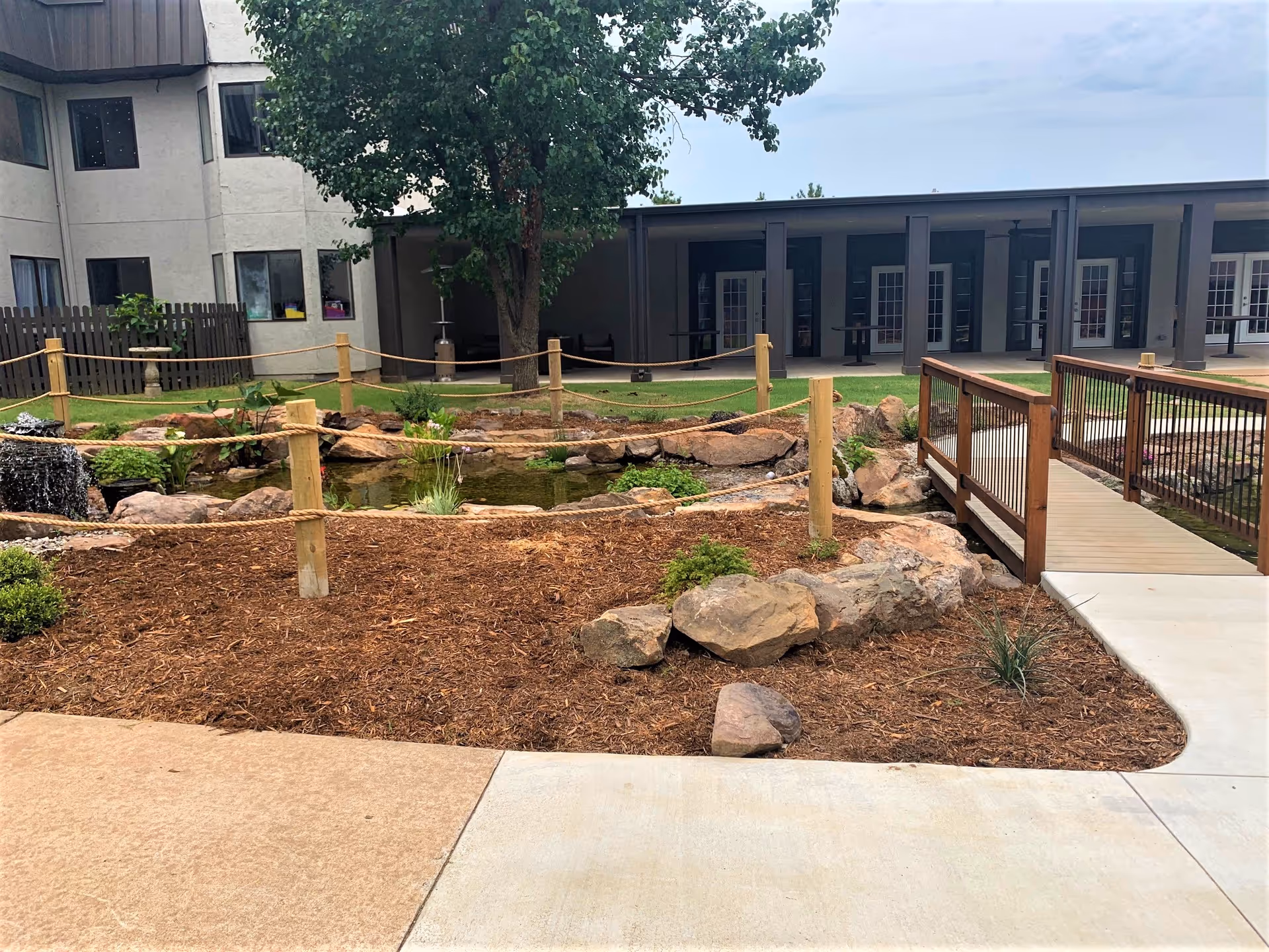 Outdoor garden area at Southern Hills Retirement Community featuring a small pond with a waterfall, surrounded by rocks and plants. There is a wooden rope fence around the pond and a wooden bridge crossing over it. In the background, there is a two-story building with multiple windows and a covered patio area with tables and chairs.